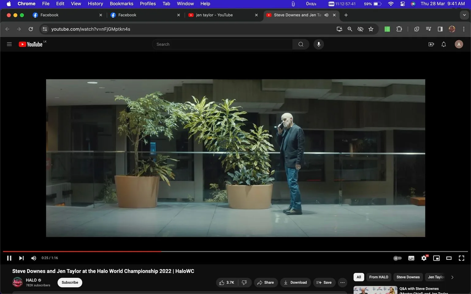 Steve Downes is featured in this screenshot from a YouTube video titled "Steve Downes and Jen Taylor at the Halo World Championship 2022 | HaloWC." The video is hosted on the HALO channel. The scene is set indoors with a modern, nighttime ambiance. Steve Downes is standing by a row of large potted plants, seemingly engaged in a phone conversation. He is dressed casually in a dark jacket, jeans, and boots, and is positioned to the right of the frame. The setting is spacious and well-lit, with a glass barrier and a muted color palette contributing to a serene atmosphere. The image conveys a moment of contemplation or communication, likely relevant to his involvement with the Halo series. The video is related to the "Halo 3" game, adding context to the interaction depicted.