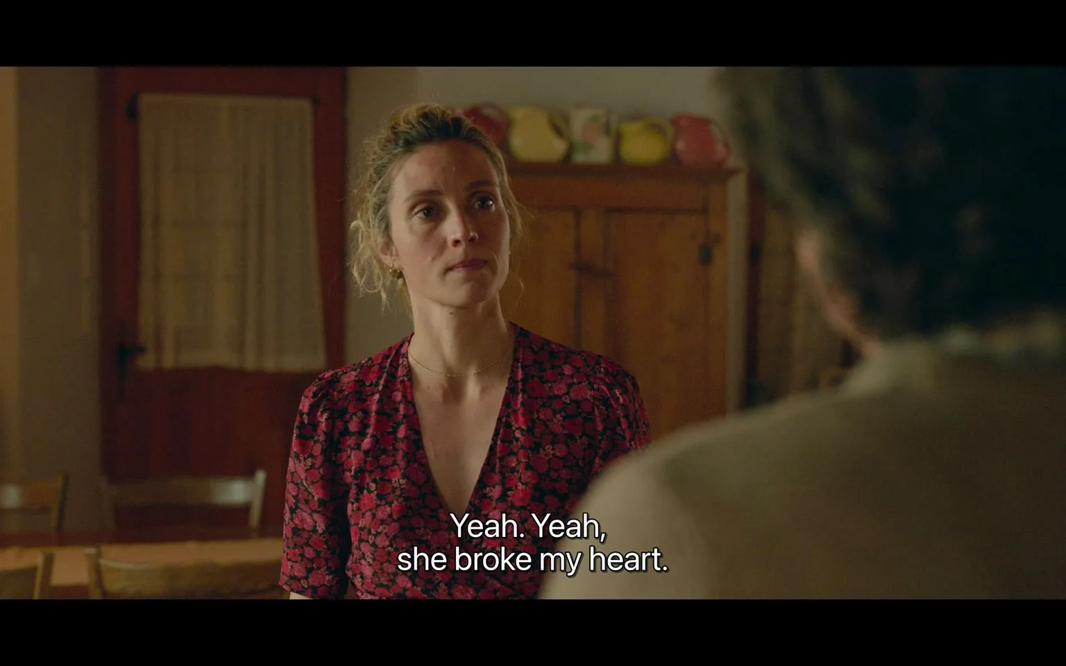 In this scene, a woman stands in a warmly lit room, engaging in an emotional conversation. She wears a red dress with a black floral pattern, and her hair is tied up casually. Her expression is serious, suggesting the conversation is meaningful or intense. The setting appears domestic, with wooden furniture and a cabinet in the background, atop which are colorful pitchers adding a homely touch. The lighting is soft, contributing to an intimate atmosphere. The subtitles reveal the emotional weight of the dialogue, with the line "Yeah. Yeah, she broke my heart," indicating a moment of vulnerability or personal revelation. The man's back is partially visible, suggesting he is listening or responding to her. The overall tone of the scene conveys themes of heartbreak and openness in a personal setting.