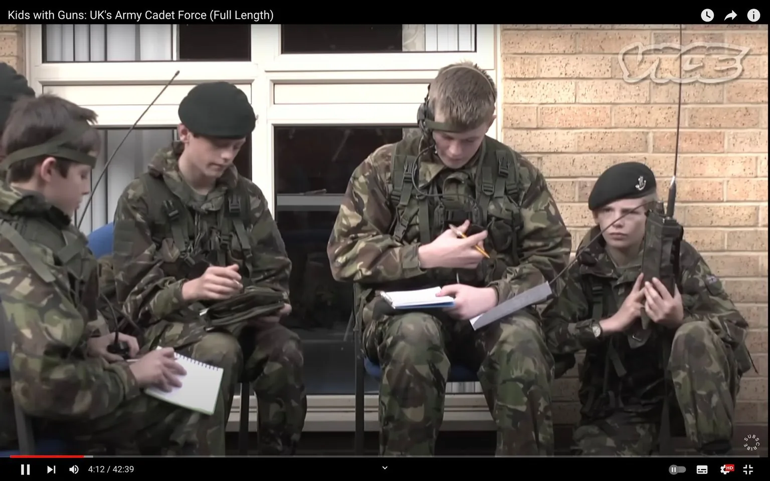 The image is a frame from a video titled "Kids with Guns: UK's Army Cadet Force (Full Length)" on YouTube, published by VICE. It shows a group of young cadets in military training. They are all dressed in camouflaged uniforms, complete with berets and various pieces of gear such as webbing and radios. The setting appears to be outdoors, possibly at a military training facility, as indicated by the brick wall and large windows in the background.
The group comprises four cadets seated and kneeling on chairs. They are engaged in an activity that involves writing or taking notes, as each holds a notepad or paper and a writing instrument. Their expressions are serious and focused, suggesting a training exercise or briefing. The lighting is natural, typical of an overcast day, which adds a neutral tone to the scene.
The cadets are arranged closely together, indicating teamwork and communication. One cadet kneels, holding a radio with a long antenna, likely involved in communication training. The overall mood of the frame suggests discipline and concentration, reflective of a structured military environment. The VICE logo is visible in the upper part of the frame, reinforcing the video's publisher.