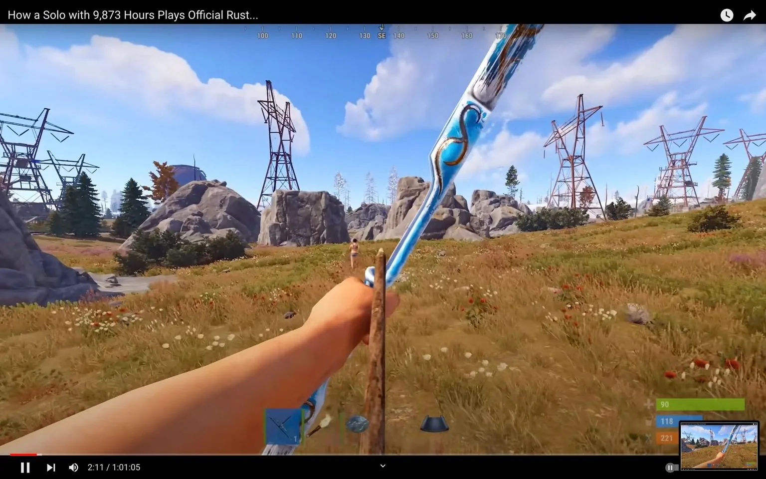This image is a frame from a gameplay video titled "How a Solo with 9,873 Hours Plays Official Rust...". The game being played is "Rust". The perspective is in first-person, showing an arm holding a bow, aimed at a distant character in a grassy field. The scene is set in an outdoor environment with a clear blue sky, dotted with a few clouds. In the distance, there are several tall structures resembling electrical towers, and rocky hills are scattered around the landscape. The player's health and inventory status are visible on the bottom right corner, showing green, blue, and orange bars. The bow is intricately designed with blue and gold patterns, suggesting a customized or unique weapon. The lush field is covered with wildflowers, adding color to the scene. The visual style is realistic, typical of the survival game genre. The on-screen text at the bottom shows the video progress as "2:11 / 1:01:05".