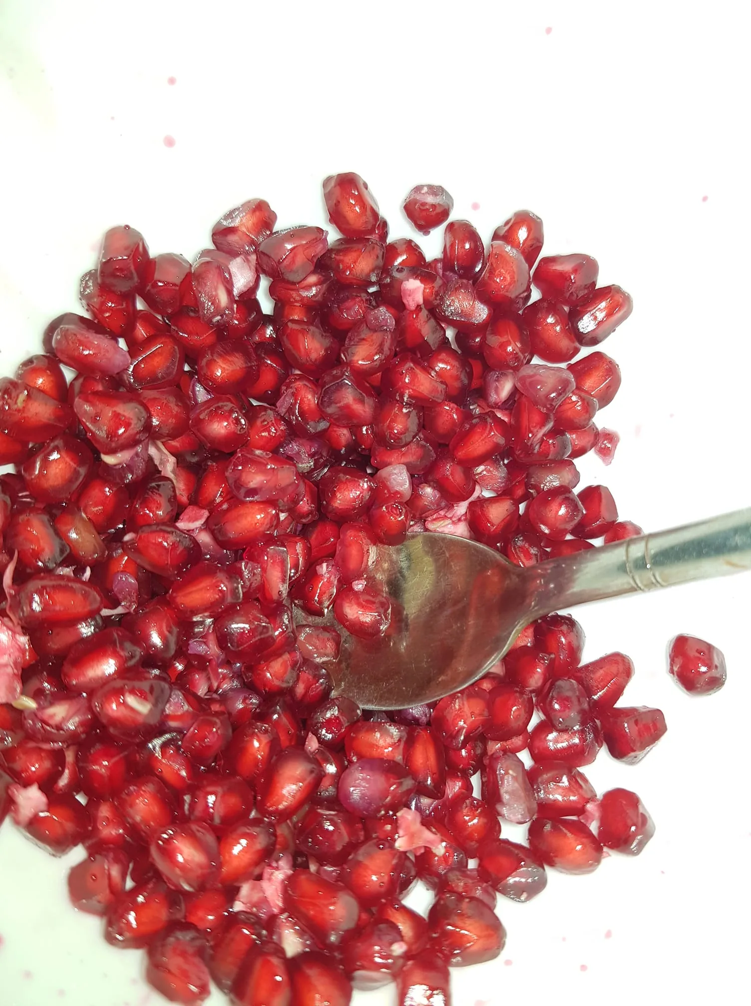 The image features a collection of vibrant red pomegranate seeds, also known as arils, scattered across a white surface. These seeds are glossy and plump, suggesting freshness and juiciness. Nestled among the seeds is a metal spoon, hinting at the idea of the seeds being ready to be eaten or served. The lighting is bright, enhancing the rich crimson color of the seeds and creating a visually appealing contrast against the white background. The focus is entirely on the pomegranate seeds, with no other objects or distractions in the frame, creating a simple yet striking composition. The mood conveyed is one of freshness and natural beauty, with the seeds likely being part of a healthy snack or meal preparation.