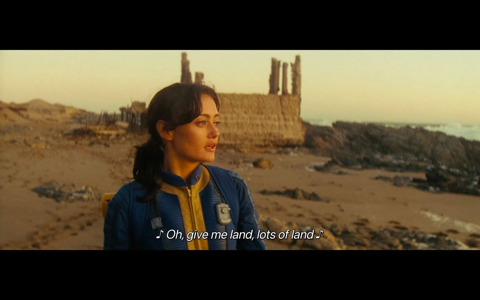 1) Rich description: The image is a screenshot from the "fallout tv show," showcasing a woman standing on a desolate beach. She is wearing a blue and yellow jumpsuit, typical of the Vault-Tec attire from the Fallout series. Her expression appears contemplative as she gazes towards the horizon. The beach is strewn with rocks and debris, and the sky is painted with warm hues, suggesting a sunset or sunrise. In the background, there's a partially ruined structure, adding to the post-apocalyptic setting typical of Fallout. The lighting is soft, casting long shadows and enhancing the serene yet somber mood of the scene. The caption at the bottom reads, "♪ Oh, give me land, lots of land ♪," indicating that she might be singing or reflecting on the vastness around her.
2) On-screen text block:
- Captions / subtitles: "♪ Oh, give me land, lots of land ♪"