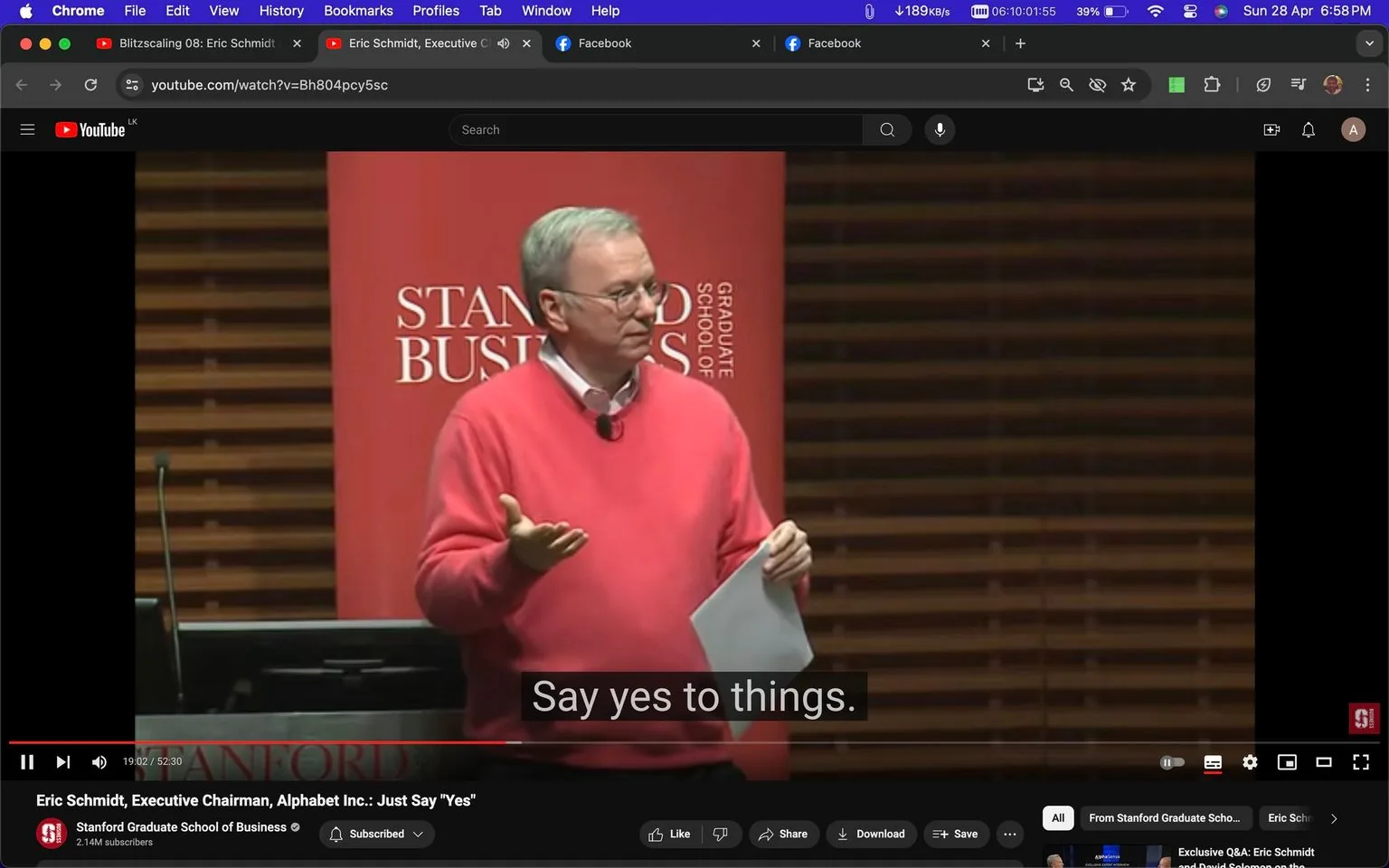 Eric Schmidt is captured mid-presentation in a lecture hall, highlighted against a backdrop featuring a red banner with the words "STANFORD BUSINESS GRADUATE SCHOOL." The setting is clearly academic, with wooden paneling suggesting a formal environment. Eric Schmidt is wearing a bright pink sweater over a collared shirt, holding a sheet of paper, and gesturing with his hand, emphasizing a point to his audience. The lighting is warm and focused on him, indicating he is the center of attention in this scene. His demeanor is engaged and confident, likely reflecting the motivational nature of his talk.
The YouTube video title is "Eric Schmidt, Executive Chairman, Alphabet Inc.: Just Say 'Yes'," and it is hosted on the Stanford Graduate School of Business channel. The subtitle "Say yes to things." appears at the bottom, capturing a key takeaway from his speech. The interface shows a Chrome browser window, with visible tabs at the top and typical YouTube controls below the video.
