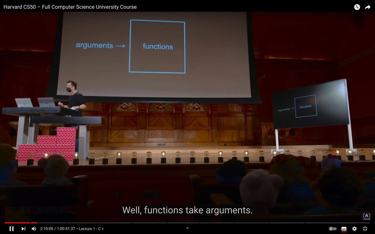 The image depicts a scene from a lecture titled "Harvard CS50 – Full Computer Science University Course," likely held in a large lecture hall. The presenter, wearing a mask, stands behind a podium with two laptops. He appears to be explaining a concept related to programming, as indicated by the slide projected on a large screen. The slide displays the text "arguments -> functions," which suggests a discussion about how functions in programming languages accept arguments. Additional monitors on stage display the same slide, ensuring visibility for the audience.
The setting is a traditional lecture hall with wooden paneling and decorative elements on the walls, creating an academic atmosphere. The lighting is warm, focusing on the presenter and the screen. The audience is visible in the foreground, sitting attentively, with some figures possibly resembling puppets due to their exaggerated features. The subtitle at the bottom of the screen reads, "Well, functions take arguments," reinforcing the educational topic being discussed. The overall composition highlights an engaging educational experience in a formal academic setting.