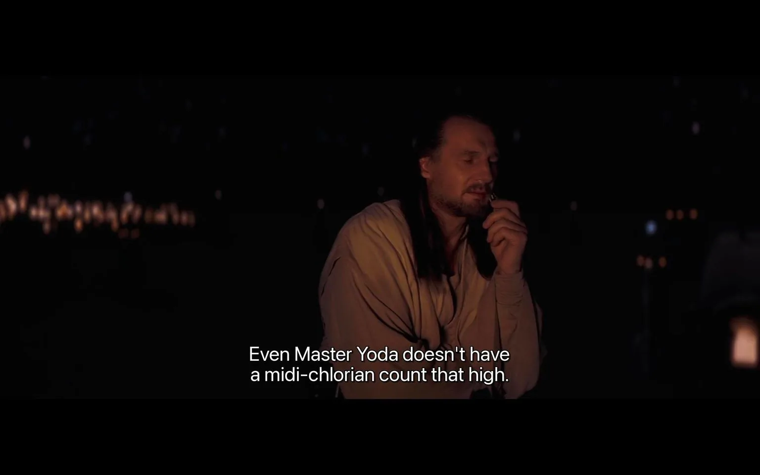The image is a night-time scene from a "Star Wars" film. It features a character in a contemplative pose, partially illuminated by what appears to be a campfire or similar light source, giving the setting a warm, ambient glow amidst the darkness. The character is dressed in traditional Jedi robes, with long hair and a thoughtful expression, suggesting a moment of reflection or discussion. In the background, small points of light suggest a distant cityscape or stars, adding depth to the scene. The dialogue at the bottom reads, "Even Master Yoda doesn't have a midi-chlorian count that high," indicating a conversation about the Force and its connection to midi-chlorians, a key concept in the "Star Wars" universe. The mood is serious and introspective, fitting the themes of destiny and power that are often explored in the series. This frame captures a moment of revelation or significance within the story.