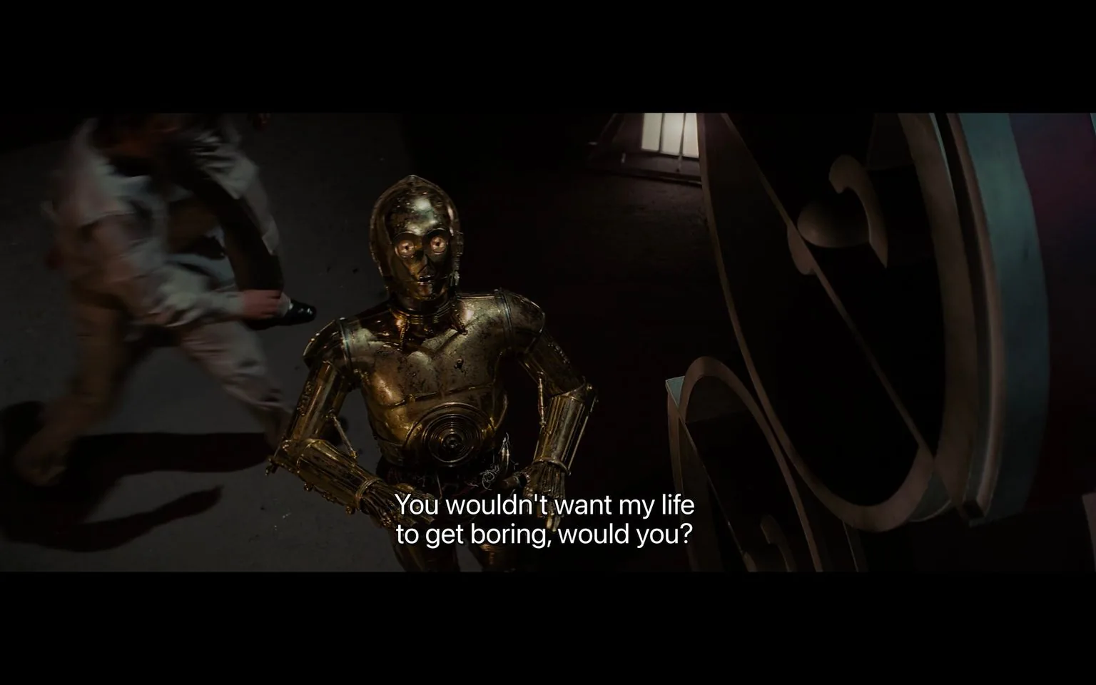 1) Rich description: The image is a screenshot from a scene in "Star Wars." It features the iconic character C-3PO, a humanoid robot with a shiny golden exterior, standing indoors. The robot's design is detailed, with visible joints and panels that reflect the light, enhancing its metallic appearance. C-3PO is looking up, possibly engaging with someone off-screen, and is positioned in the center foreground. The background is dark, with a slightly blurred figure moving past, adding a sense of motion and urgency to the scene. The lighting is soft yet dramatic, highlighting the robot's features against the shadowy setting. The subtitle, "You wouldn't want my life to get boring, would you?" suggests a whimsical or humorous tone in C-3PO's dialogue. The environment appears industrial, with structures that have an angular and mechanical aesthetic, consistent with the "Star Wars" universe's futuristic theme.
2) On-screen text block:
- Captions / subtitles: "You wouldn't want my life to get boring, would you?"