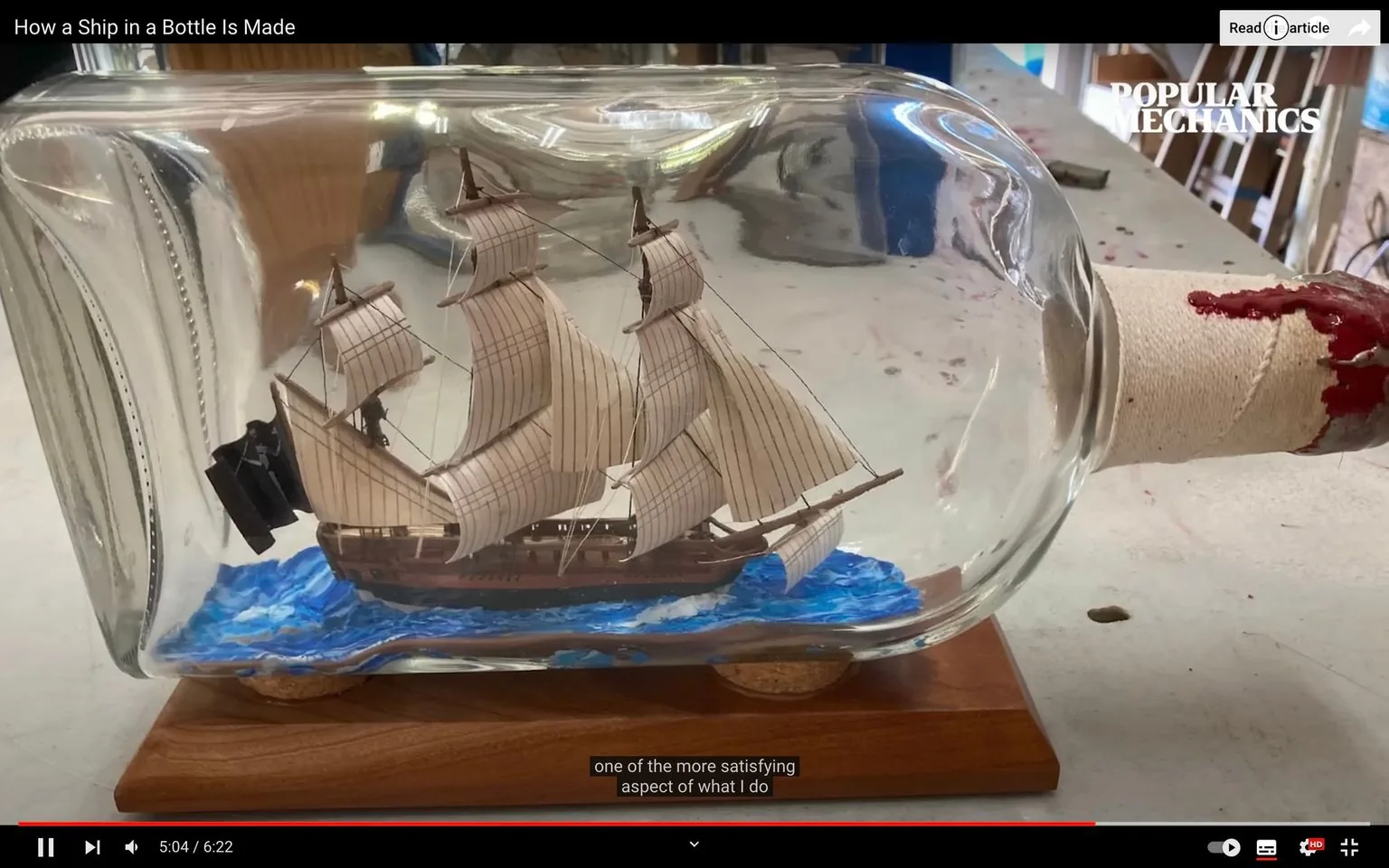 This image is a still from a video titled "How a Ship in a Bottle Is Made," featured on YouTube by the channel Popular Mechanics. The scene shows a detailed model of a sailing ship inside a glass bottle. The ship is intricately crafted with tan sails and a brown wooden hull, set against a backdrop of painted blue waves. The bottle is positioned on a wooden stand, and the neck of the bottle is wrapped with a cloth sealed with red wax. The setting suggests a workshop environment, indicated by the tools and surfaces visible in the background. The lighting is bright, highlighting the craftsmanship of the model. The subtitle at the bottom reads, "one of the more satisfying aspect of what I do." The video explores the intricate process of creating these miniature marvels, showcasing the skill and patience involved in this traditional craft. The Popular Mechanics logo is visible in the upper right corner, reinforcing the channel’s focus on detailed and technical craftsmanship content.