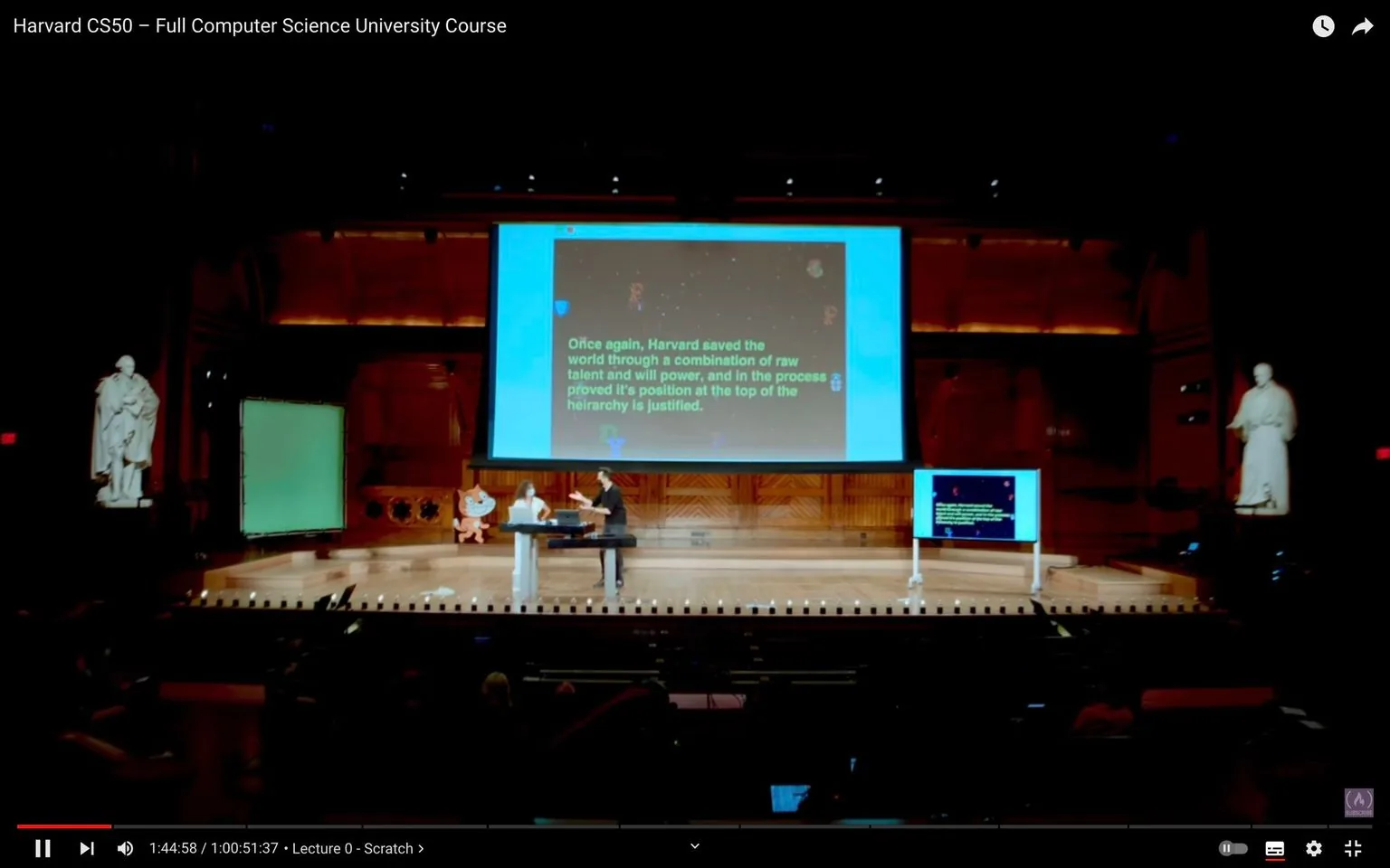 The image depicts a scene from a Harvard CS50 lecture, specifically the "Lecture 0 - Scratch" segment. The setting is a large lecture hall with a wooden interior, reflecting a traditional academic atmosphere. At the center is a sizable screen displaying text, and a smaller monitor to the side mirrors this content for better visibility. The stage features two individuals, likely instructors or presenters, engaged in a discussion or demonstration. The text on the screen humorously acknowledges Harvard's reputation, stating, "Once again, Harvard saved the world through a combination of raw talent and will power, and in the process proved it's position at the top of the hierarchy is justified."
The setting is illuminated by warm lighting, and the hall is spacious with statues flanking the stage, adding a scholarly touch. The audience can be faintly seen in the foreground, indicating the lecture is well-attended. The atmosphere is academic and engaging, with the focus on the presentation and teaching elements typical of a university course. The video is part of Harvard's educational content available on YouTube, showcasing their popular computer science course.