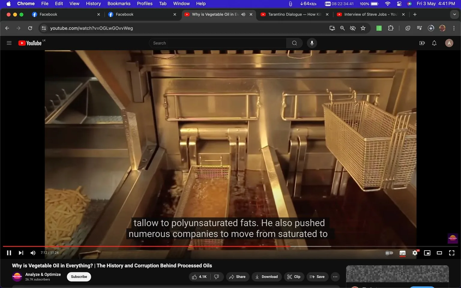 The image is a screenshot from a YouTube video titled "Why is Vegetable Oil in Everything? | The History and Corruption Behind Processed Oils," presented by the channel "Analyze & Optimize." The scene shows a commercial deep fryer with multiple baskets, some of which contain French fries, submerged in hot oil. The fryer is made of stainless steel, suggesting a professional kitchen setting. Above the fryer, subtitles overlay the video, discussing the transition of fats from tallow to polyunsaturated oils, indicative of the video's theme focusing on dietary fats and industry practices. The lighting is bright, highlighting the shiny metal surfaces and bubbling oil. The browser tabs at the top indicate that this scene is watched in a Chrome browser, with other tabs open for Facebook and additional YouTube videos, suggesting multitasking. The video's progress bar shows it is 7 minutes and 12 seconds into an 11-minute and 24-second video. On-screen icons like thumbs up, share, and save are visible, indicating user interaction options.
