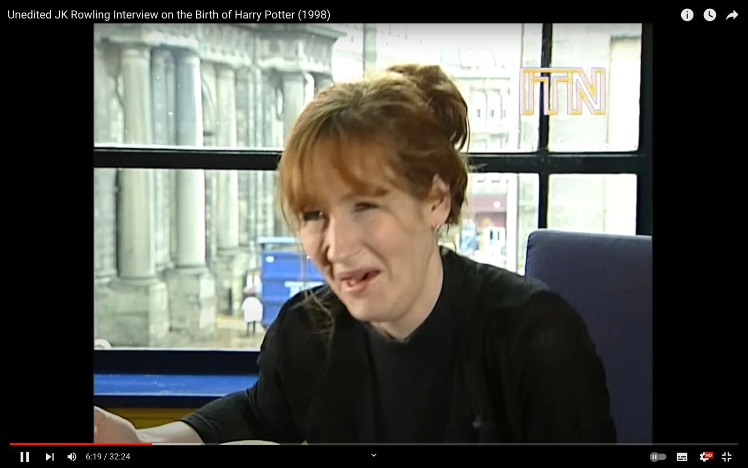 1) Rich description: The image is a video frame from an interview with JK Rowling in 1998, discussing the birth of "Harry Potter." The setting is indoors, with a large window in the background revealing blurred urban architecture, suggesting a cityscape. The window frames the subject, JK Rowling, who is seated and facing the camera. She is captured in a moment of laughter, with her head slightly tilted and a warm expression on her face. Rowling's hair is auburn and styled in a loose bun, and she is wearing a black top and cardigan, which contrast with the light coming through the window. The lighting in the room is natural, highlighting her features softly. The ITN logo is visible in the top right corner, indicating the broadcaster. The overall tone of the image is casual and intimate, capturing a moment of candid conversation.
2) On-screen text block:
- Title: "Unedited JK Rowling Interview on the Birth of Harry Potter (1998)"
- Publisher / studio: "ITN"