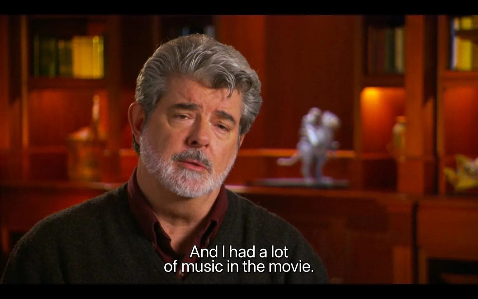 George Lucas is shown in an interview setting, seated against a backdrop of a warm, wood-paneled library. The shelves behind him are filled with books and softly lit ornamental objects, creating a cozy and intellectual ambiance. George Lucas appears to be speaking thoughtfully, with a calm expression and a slight tilt of his head. He is wearing a dark sweater over a maroon shirt, which adds to the professional yet relaxed atmosphere. The lighting is soft and focused on him, highlighting his face and adding depth to the scene. The caption at the bottom of the image reads, "And I had a lot of music in the movie," suggesting he is discussing his experience with film music. The overall composition centers on Lucas, emphasizing his presence and words in the image.