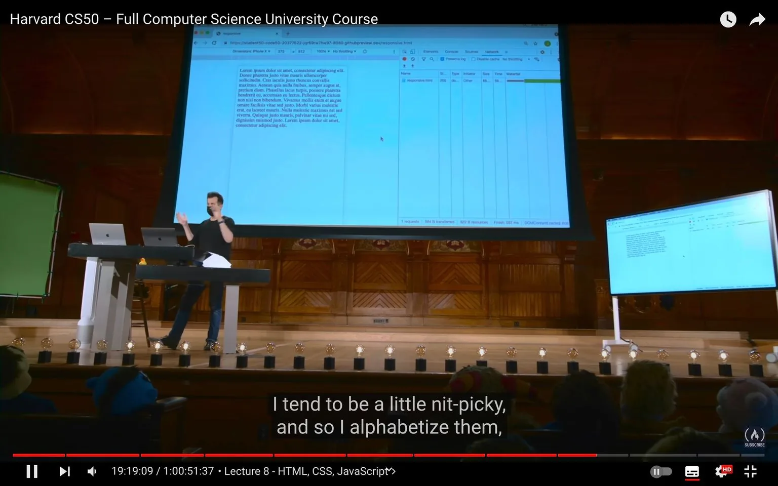 The image captures a scene from the Harvard CS50 course, specifically during lecture 8, which covers HTML, CSS, and JavaScript. The setting is a large lecture hall with a wooden backdrop, where an instructor is actively presenting. He stands at a lectern, gesturing as he explains concepts displayed on a large screen behind him. The screen shows a web browser window with developer tools open, displaying a network tab alongside some placeholder text.
The instructor is casually dressed in a dark shirt and jeans, creating a relaxed yet focused atmosphere. Several laptops are on the lectern, indicating the use of technology in the presentation. On the stage, a series of small lights are positioned along the edge, adding to the professional setup. The audience, visible in the foreground, appears engaged with the lecture.
Below the video frame, subtitles are provided, capturing a line where the instructor mentions being “nit-picky” and his habit of alphabetizing. This adds a personal touch to the technical content being discussed. The video's title at the top reads "Harvard CS50 – Full Computer Science University Course," indicating the educational nature of the content.
