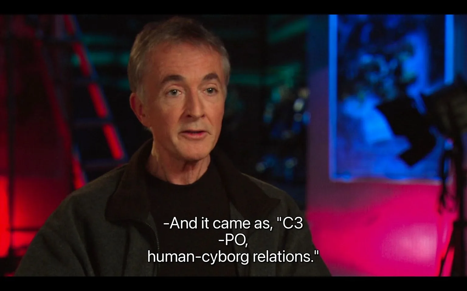 In this video frame, an interview is taking place in a studio setting. The person, who is not identified by name, is speaking about the character C-3PO from "Star Wars." The background features vibrant, colorful lighting with shades of red and blue, creating a visually striking contrast against the darker, more subdued clothing of the speaker. The individual is dressed in a dark jacket and black shirt, and their expression is attentive and engaged, suggesting a thoughtful discussion. The framing places the speaker slightly off-center, emphasizing the colorful backdrop that may include studio equipment and indistinct shapes adding to the professional setting. The subtitle indicates a quote related to the character's introduction: "'C3 -PO, human-cyborg relations.'"
