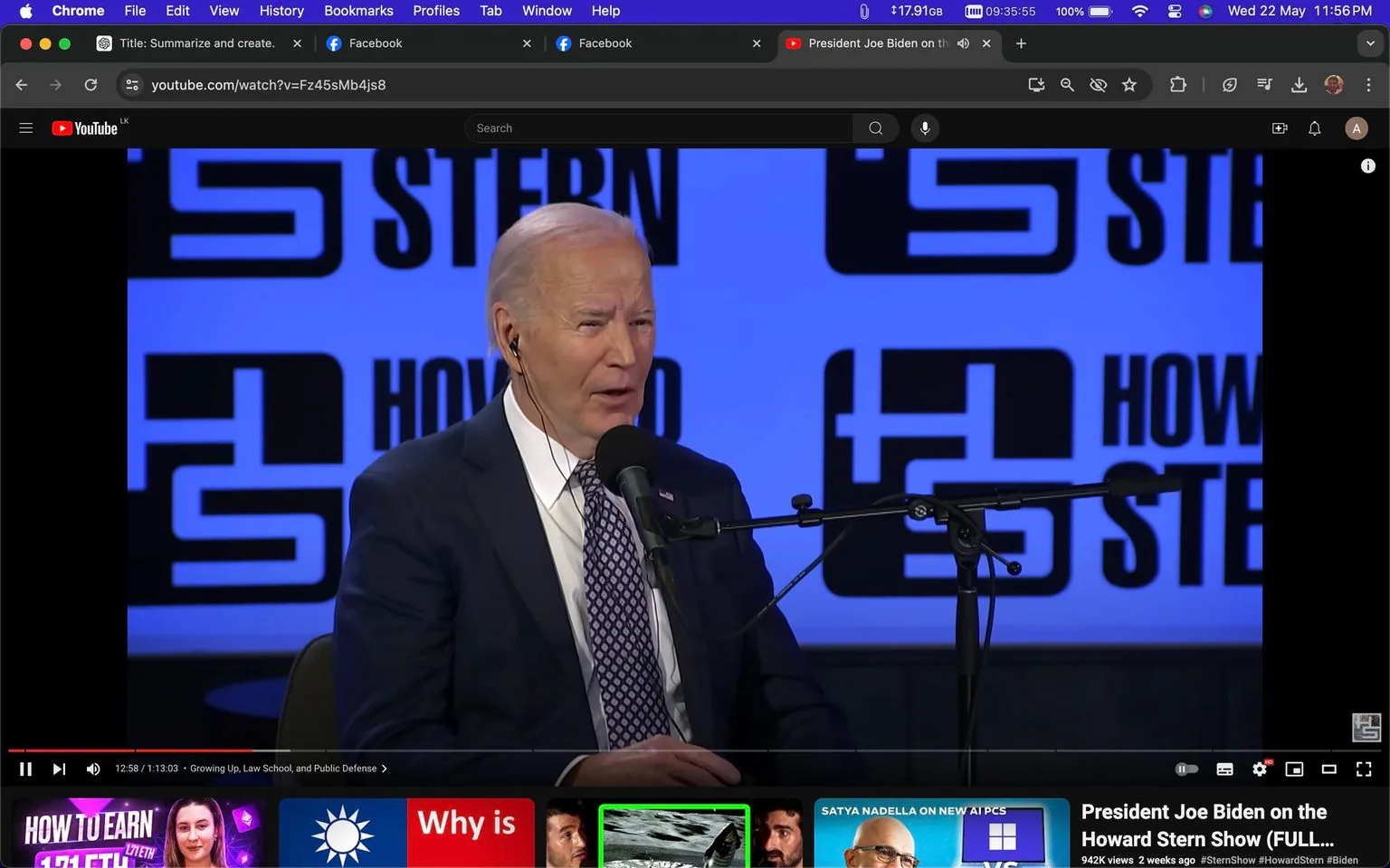 In this screenshot, President Joe Biden is featured during an interview on the Howard Stern Show. The setting is a studio environment, characterized by a prominent blue backdrop displaying the words "STERN" and "HOWARD" in a bold, modern font. Joe Biden is seated in front of a microphone, wearing a dark suit with a white shirt and a patterned tie, corresponding to a formal appearance. He has an earpiece in his left ear, indicating an ongoing conversation or broadcast. The lighting is bright and focused, highlighting Biden's expressions as he appears to be speaking. The YouTube interface is visible, with a video title partially reading "President Joe Biden on the Howard Stern Show (FULL..." and a timestamp indicating the video progress at 12 minutes and 58 seconds of a 1 hour and 13-minute video. Below the main video, there are thumbnails of other suggested videos, including topics related to technology and personalities. The presence of the YouTube logo and typical interface elements like the search bar and user icon are also visible, solidifying the platform as YouTube.