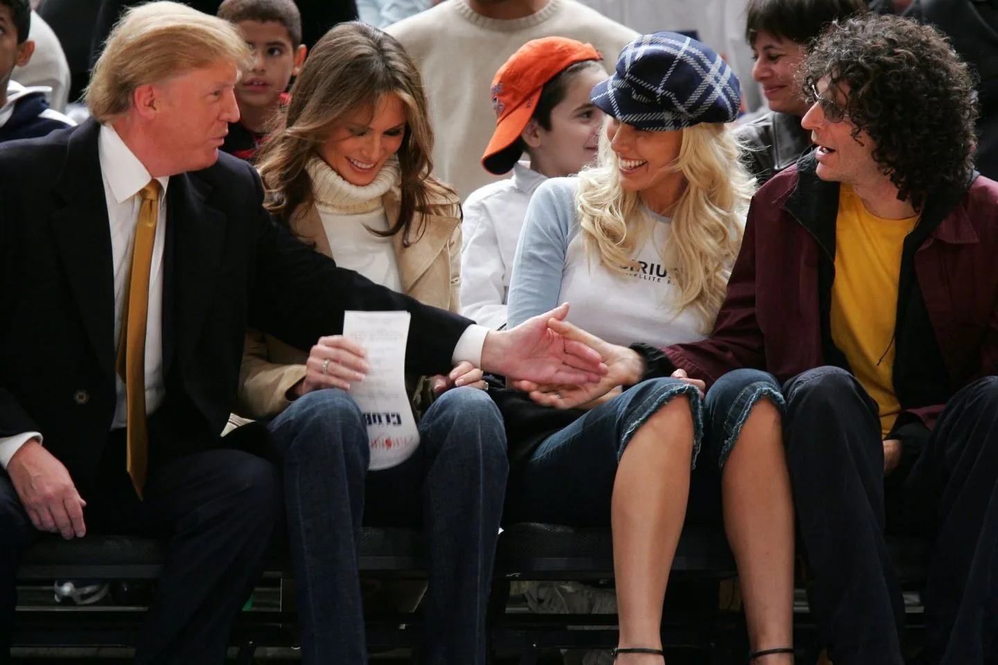 In this image, we see a group of people seated closely together, likely at a public event such as a sports game. The setting suggests an indoor arena with several spectators visible in the background, indicating a lively and engaging atmosphere. On the left, a man in a suit with a gold tie is reaching over to shake hands with a woman seated next to him. The woman, wearing a light-colored turtleneck sweater and a tan coat, is smiling and appears to be holding a paper. Next to her is another woman with long blonde hair wearing a checkered hat, a casual shirt, and jeans, also smiling, suggesting a friendly interaction. On the far right is Howard Stern, identified by the hint, wearing glasses, a yellow shirt, and a dark jacket, engaged in the conversation with the group. The lighting is bright, typical of indoor venues, and the overall mood is casual and social. There are no visible signs or text in the image to provide additional context.