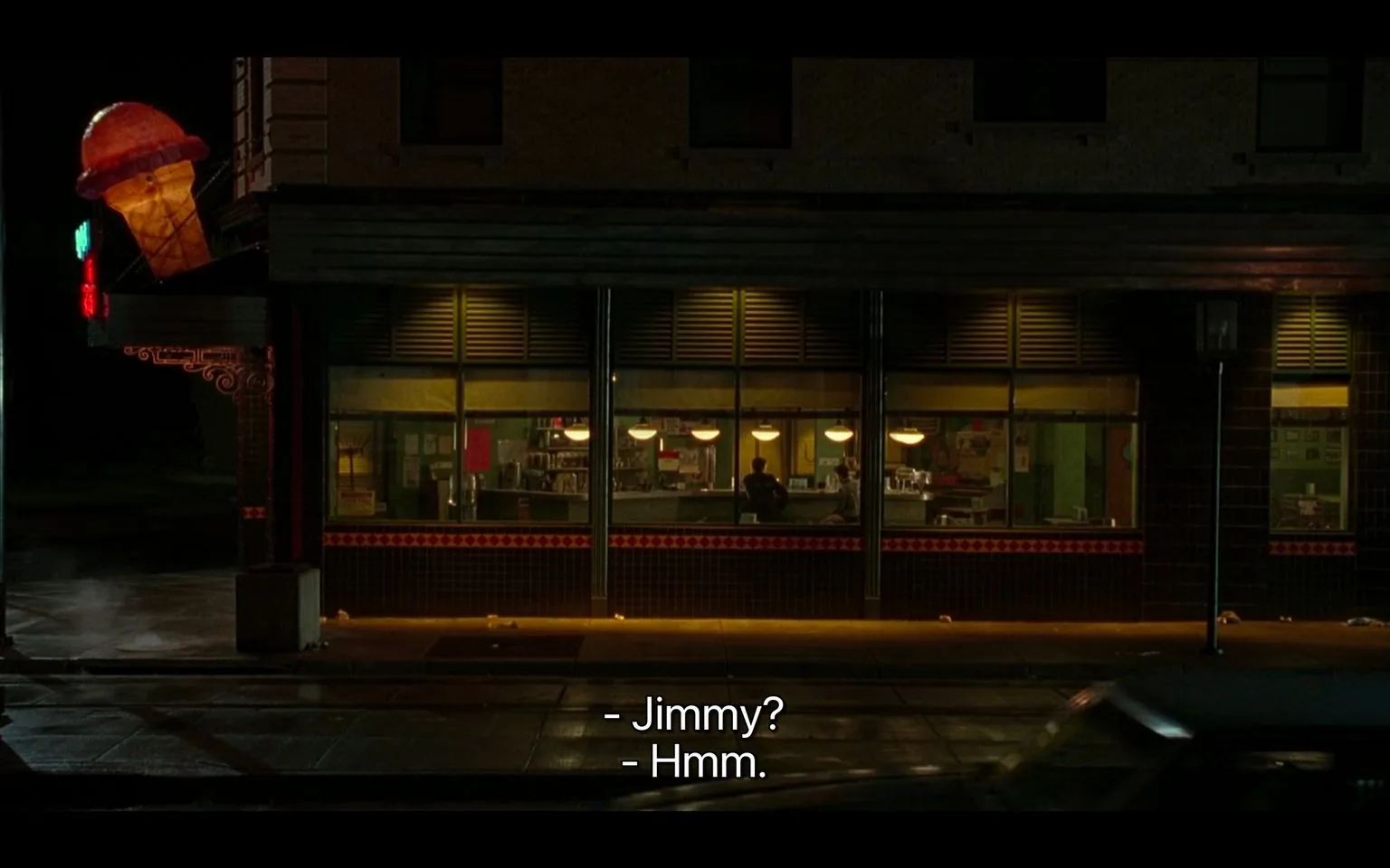 The image depicts a nighttime scene outside a diner with large windows, revealing a glimpse into the warmly lit interior. The exterior is marked by a prominent, illuminated ice cream cone sign, suggesting the establishment might serve desserts or ice cream. The street is wet, likely after rain, reflecting the ambient lights and adding a moody atmosphere. Inside, two figures can be seen sitting at the counter, engaged in conversation. The warm glow from the diner contrasts with the dark street, creating a cozy, intimate setting. The interior is decorated with various posters and signs on the walls, and several pendant lights hang from the ceiling, contributing to the homely atmosphere. The dialogue captured in the subtitles, “- Jimmy? - Hmm.”, indicates a casual conversation, suggesting familiarity between the speakers. The presence of a car in motion at the edge of the frame adds a sense of life and movement to the otherwise quiet scene.