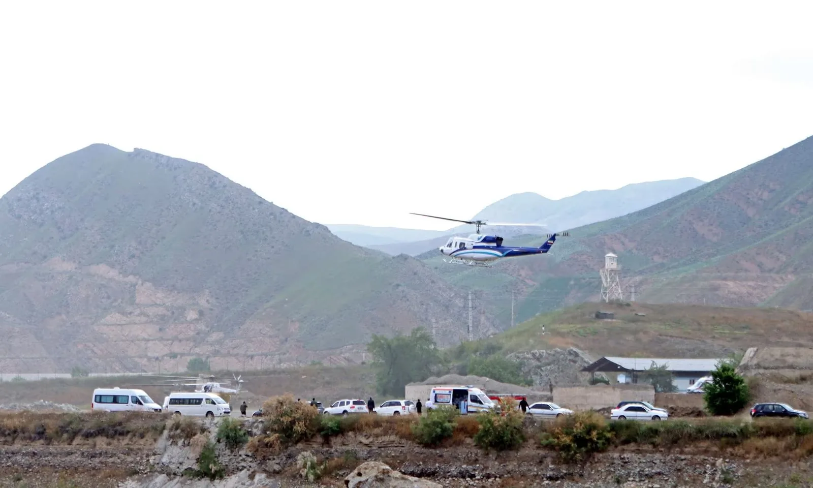 1) Rich description: The image depicts an outdoor scene with a helicopter in mid-air, flying over a landscape of rolling hills and mountains. In the foreground, there are several white vehicles, including vans and cars, parked on a dirt road. The helicopter is painted in blue and white, contrasting against the muted greens and browns of the mountainous backdrop. A group of people can be seen near the vehicles, possibly engaged in some coordinated activity. The lighting is natural, suggesting daytime, though the sky appears overcast, casting a diffuse light over the scene. A watchtower is visible in the distance, adding an element of security or surveillance. The terrain is rugged, with sparse vegetation, typical of a rural or remote area.
2) On-screen text block:
- Key OCR Lines: "Alex Marsh", "Jonathan"
