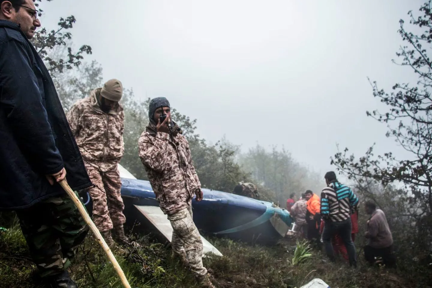 The image depicts an outdoor scene in a foggy forest area. In the foreground, several individuals are gathered around what appears to be the wreckage of a small aircraft. Two of the people are wearing camouflage clothing, and one is speaking into a walkie-talkie, suggesting a rescue or recovery operation. The presence of fog and the dense tree line indicate a secluded and challenging environment. Other individuals are dressed in various outdoor gear, including a striped jacket and a bright orange jacket, further emphasizing a coordinated effort. The mood seems tense and focused, likely due to the circumstances surrounding the crash. The image captures the urgency and seriousness of the response to the incident.
- Captions / subtitles: "..."
- Key OCR Lines: "I have to launch off our website soon", "But we have to hide too much stuff", "I have to get armed security", "Alex Marsh:", "I want my friends to own my business", "I'm touched you want to donate", "And Jonathan wants to donate volunteer hours", "But I have to sell equity out or hand it over", "I prefer it"