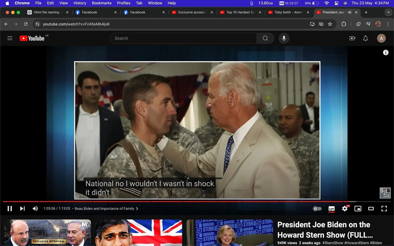 The image is a video frame from a YouTube video titled "President Joe Biden on the Howard Stern Show (FULL...)." The video is hosted on the Howard Stern channel. In the scene, two men are engaged in a conversation indoors. One man is wearing a light-colored suit with a tie, while the other is dressed in a military uniform. The man in the suit is placing his hand on the shoulder of the man in uniform, suggesting a moment of connection or support. The background shows a couple of other men in military attire, and patriotic decorations are visible, hinting at an official or ceremonial setting. The lighting is bright, emphasizing the significance of the interaction. Subtitles on the screen read, "National no I wouldn't I wasn't in shock it didn't." The video frame includes topic tags such as "#SternShow," "#HowardStern," and "#Biden," indicating the content's focus on the personalities involved.