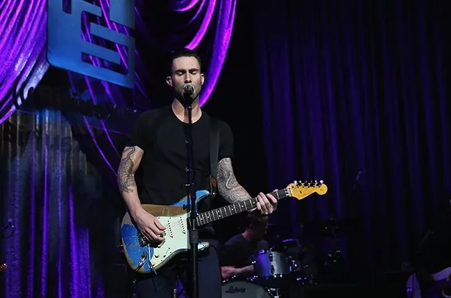 Adam Levine is performing on stage, holding a guitar and singing into a microphone. The setting is a concert venue with a dark and intimate atmosphere, enhanced by the deep purple lighting and elegant drapery in the background. Adam Levine is dressed in a simple black t-shirt, accentuating his tattooed arms, which are visible as he plays the guitar. The guitar is prominently colored with a blue body and white pickguard, adding a splash of vibrancy to the scene. Behind him, a drummer is partially visible, contributing to the live performance ambiance. The overall mood is engaging and dynamic, capturing the energy of a live musical performance. The lighting highlights Adam Levine, casting shadows that emphasize the contours of the stage setup and curtain backdrop. No visible text or branding is present that adds context to the performance.