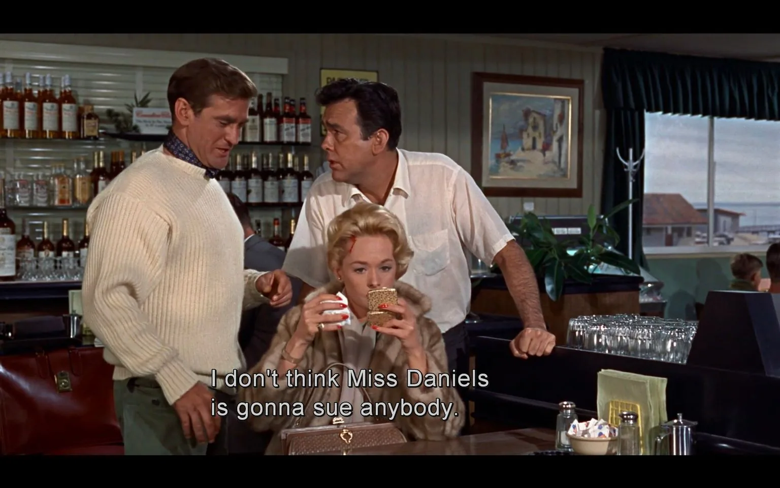 In this scene from the film "The Birds," three characters are gathered inside a bar, engaged in a conversation. The setting is cozy and atmospheric, with bottles of liquor neatly arranged on shelves along the wall in the background, suggesting a well-stocked bar. The lighting is warm, highlighting the retro decor and a painting hanging on the wall, which features a seaside village scene.
The man on the left is dressed in a cream-colored sweater and appears to be speaking to the woman seated at the bar. He is leaning slightly forward, indicating engagement in the conversation. The woman, referred to as Miss Daniels in the subtitles, is sipping a drink from a cup while holding a compact mirror, suggesting a mix of casual and attentive demeanor. She has a small cut on her forehead, adding an element of intrigue or past tension. Her blonde hair is styled in a typical 1960s fashion, and she wears a fur coat, hinting at sophistication or affluence.
Another man stands to the right of Miss Daniels, wearing a white shirt, further adding to the sense of an informal yet involved discussion. The subtitle reads, "I don't think Miss Daniels is gonna sue anybody," indicating a conversation about a potential dispute or incident, perhaps related to the events unfolding in the film. The scene captures a moment of interpersonal dynamics amidst the unfolding drama of "The Birds."