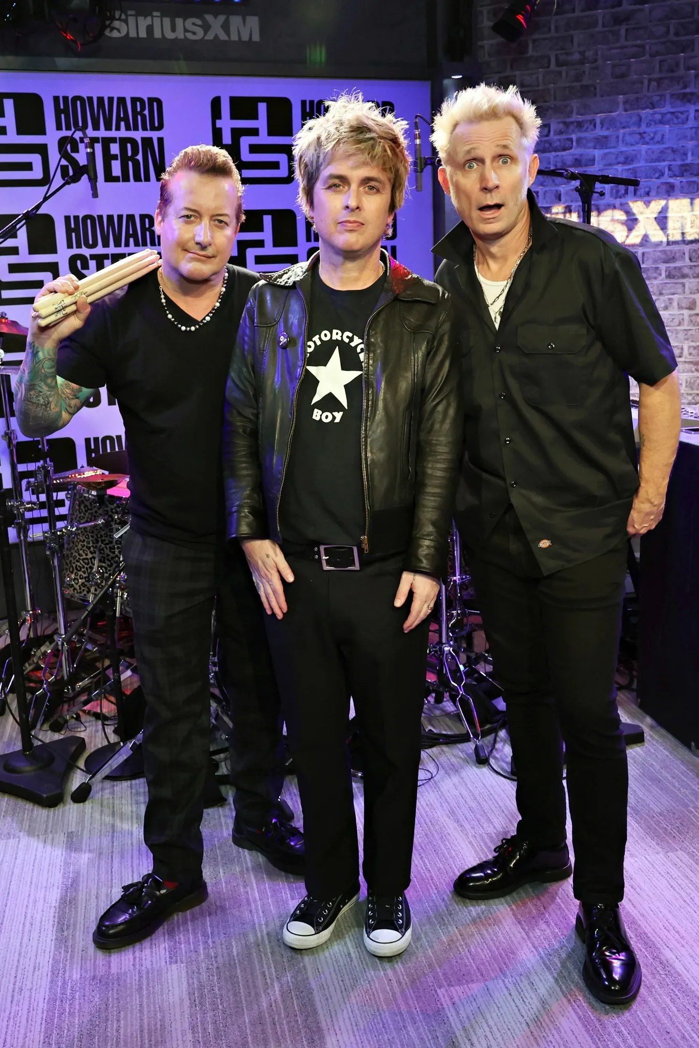 The image features the band Green Day during an appearance on "The Howard Stern Show" at the SiriusXM studio. The setting appears to be a music studio, with a drum set visible in the background, indicating a possible performance or interview setting. The background is adorned with repeated logos of "HOWARD STERN" and "SiriusXM," establishing the location and context of the scene.
The three band members are standing side by side. The person on the left is holding drumsticks, suggesting his role as the drummer. He is wearing a black t-shirt and has a tattoo visible on his arm. The central figure is wearing a black leather jacket over a t-shirt with the words "MOTORCYCLE BOY" and a star design, giving a rock-and-roll vibe. The person on the right is wearing a black shirt and has a surprised or animated expression on his face. The studio lighting is bright, casting a clear view of the band members, with a mix of casual and performance attire. The mood is relaxed yet energetic, fitting the band's punk rock image.