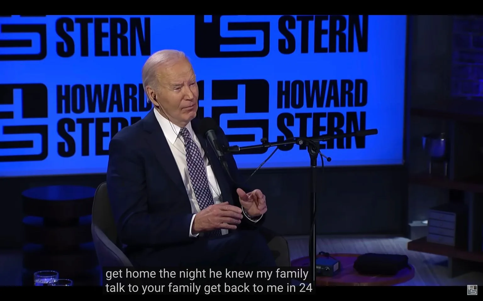 1) Rich description: The image depicts a scene from an interview setting, featuring Biden speaking into a microphone. He is seated in a studio environment with a prominent blue background displaying the text "HOWARD STERN" repeatedly in bold, black letters. Biden is dressed in a formal dark suit, white shirt, and patterned tie. He appears to be engaged in conversation, with a thoughtful expression, gesturing slightly with his hands. The studio setup includes professional audio equipment with a microphone positioned in front of him. A small table nearby holds a couple of glasses, suggesting a casual and conversational atmosphere. The lighting is bright, and the overall ambience of the studio is modern and sleek, emphasizing a professional interview setting.
2) On-screen text block:
- Captions / subtitles: "get home the night he knew my family talk to your family get back to me in 24"