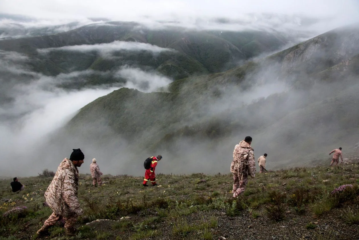 The image depicts a group of people walking down a misty, mountainous landscape. In the foreground, several individuals are dressed in camouflage clothing, suggesting they might be part of a search or rescue team. Among them, one person stands out in a bright red and yellow uniform with a backpack, indicating they might be a specialized member of the team possibly involved in emergency or rescue operations. The mood is serious and focused as they navigate the steep, grassy terrain.
In the background, layers of rolling mountains are partially obscured by dense fog, creating a mysterious and serene atmosphere. The early morning or late afternoon light adds a muted gray tone to the scene, enhancing the dramatic effect of the mist. The overall composition emphasizes the vastness and ruggedness of the natural environment, with the rescue team's effort to traverse it forming the central focus. The image captures a moment of action and determination against a backdrop of natural beauty and challenge.