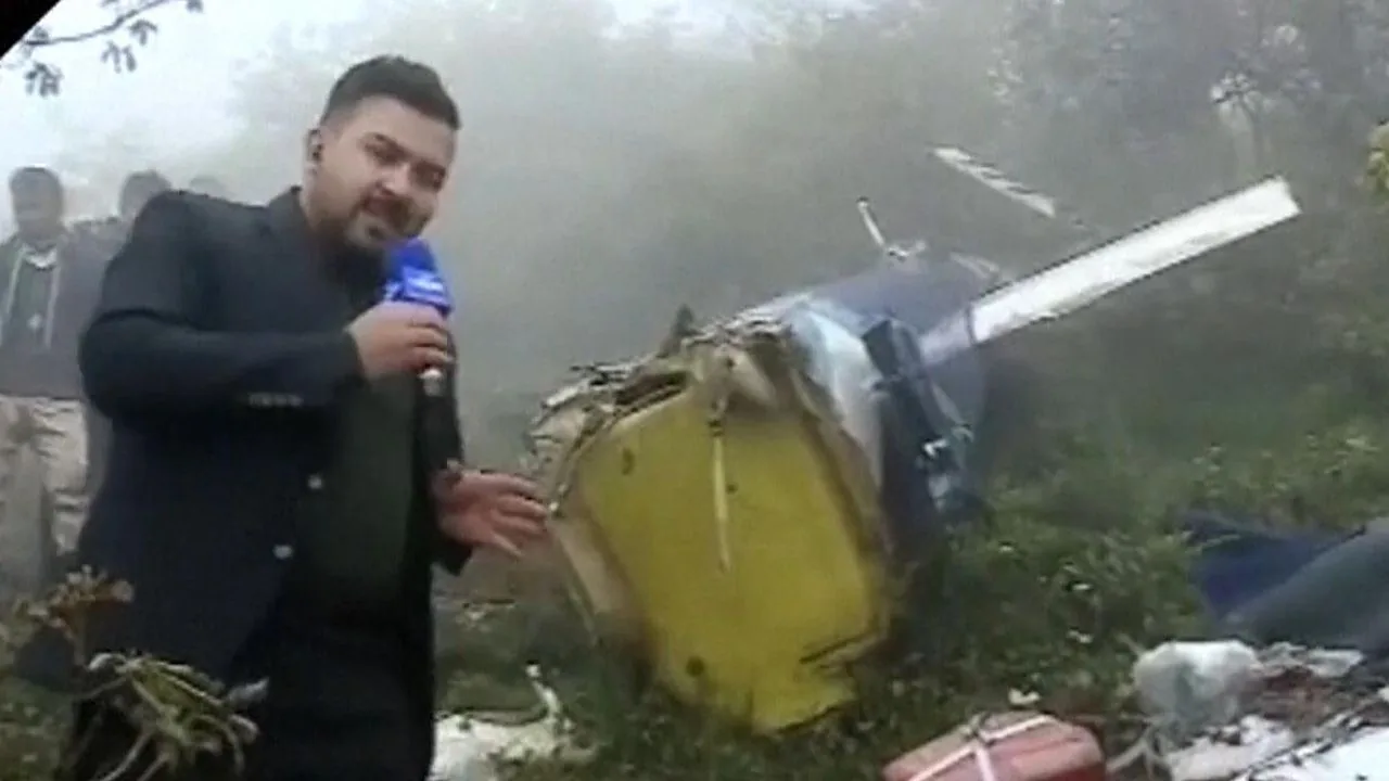 The image depicts a reporter at what appears to be a crash site. The environment is misty, with fog obscuring the background, indicating it might be early morning or a cloudy day. The reporter is holding a microphone, suggesting they are delivering a live report or recording a news segment. The wreckage of a small aircraft or helicopter is visible beside him, partially covered by grass and debris. The reporter is dressed in a dark suit, adding a formal tone to the scene. The mood is serious, as expected in a report involving an accident. Surrounding the reporter, a few individuals are visible, possibly investigators or onlookers. The colors are muted due to the foggy conditions, and the overall atmosphere is somber and tense, highlighting the gravity of the situation. There is no visible branding or text on the microphone that identifies the news organization.