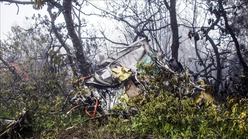 The image depicts a scene of aircraft wreckage amidst a forested area. The debris is tangled with the surrounding trees and vegetation, indicating a severe impact. Various pieces of metal and components are visible, some identifiable as parts of an aircraft structure. The setting is dense with trees, and there is a misty or foggy atmosphere, adding to the somber mood of the scene. The foreground shows damaged and twisted metal, with parts colored in muted tones, primarily gray and silver, with occasional hints of color from wires or fabric. The background is filled with trees, some with leaves and others barren, hinting at possible seasonal change. The lighting is dim, consistent with overcast weather or the aftermath of a fire. There are no visible signs of fire or smoke damage on the wreckage itself, but the overall scene suggests a recent or past incident.