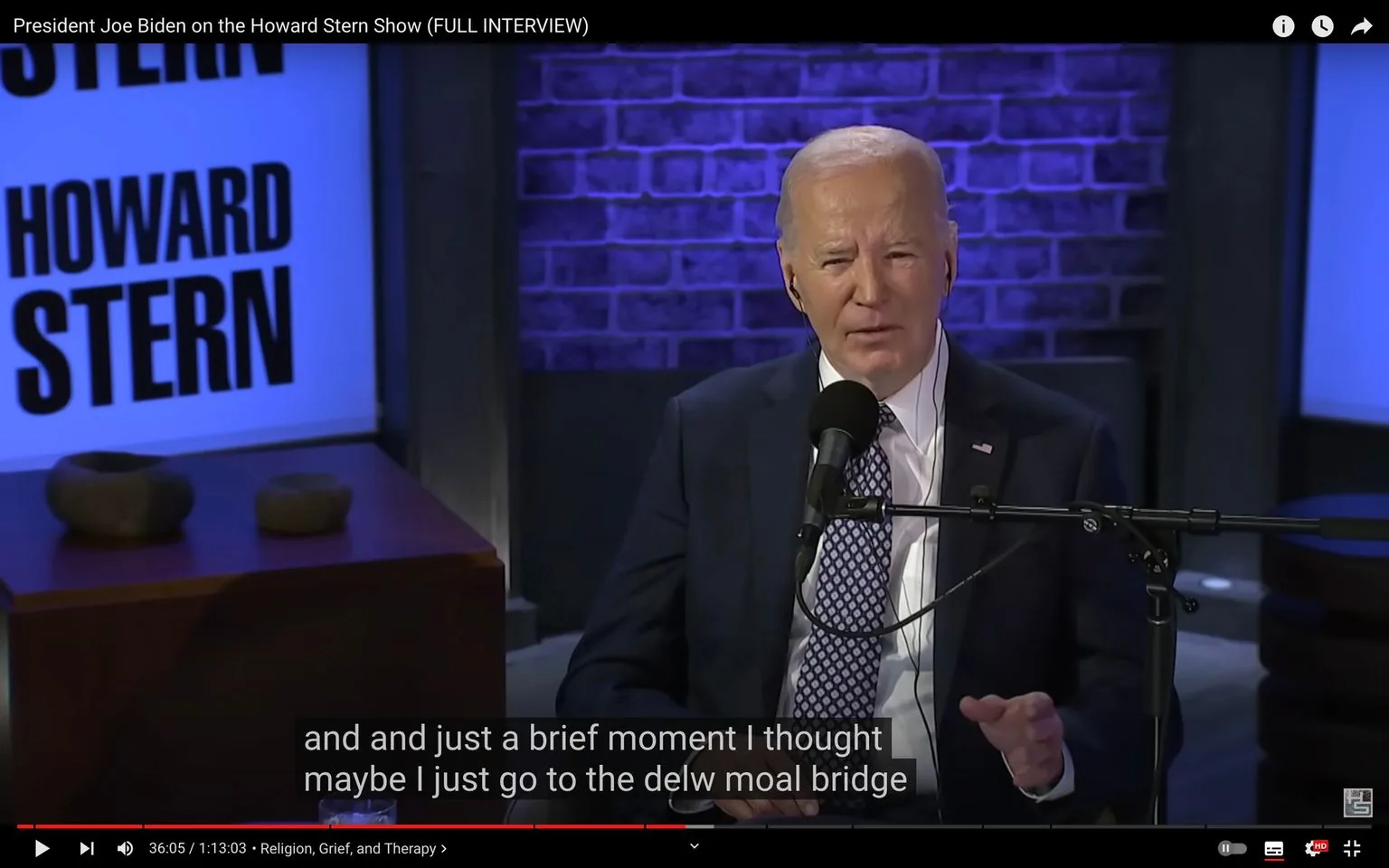 1) Rich description: The image is a video frame from an interview featuring Joe Biden on the "Howard Stern Show," as indicated by the hint and title text. Biden is seated in a studio setting with a microphone in front of him, suggesting he is engaged in a conversation or interview. He is dressed in a formal suit and tie, appearing serious and focused. The background features a brick wall with purple lighting, creating a professional yet casual atmosphere typical of talk shows. To the left, a sign with the text "HOWARD STERN" is prominently displayed, confirming the show's setting. The video frame includes subtitles, currently displaying Biden speaking about a brief moment of contemplation. The scene is indoors, with a polished, media-ready setup typical of high-profile interviews. The lighting is even, highlighting Biden and ensuring clarity for viewers.
2) On-screen text block:
- Title: "President Joe Biden on the Howard Stern Show (FULL INTERVIEW)"
- Captions / subtitles: "and and just a brief moment I thought maybe I just go to the delw moal bridge"