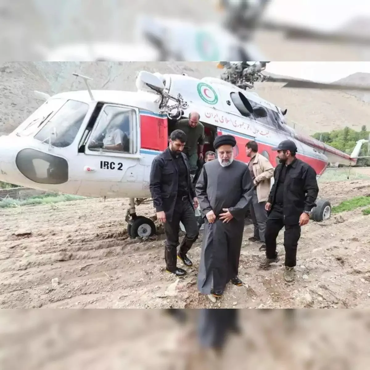 The image captures a group of people disembarking from a helicopter in a rugged, outdoor setting. A notable figure, dressed in traditional attire, including a black robe and hat, is accompanied by several other men in dark suits, possibly security personnel. The presence of a helicopter with visible text "IRC 2" suggests this might be an official or humanitarian visit. The helicopter is white with a red stripe and has markings in another language, possibly identifying it as part of a specific organization. The landscape is mountainous and appears remote, with a dirt ground underfoot. The mood seems formal and purposeful, as the group steps away from the helicopter towards an unspecified destination.