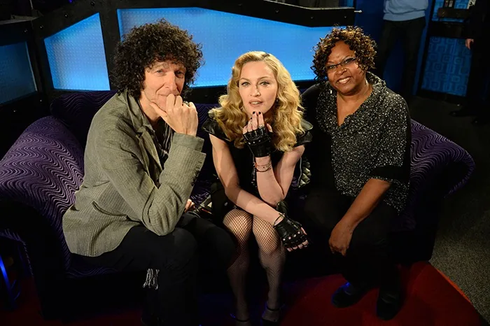 The image captures a studio setting with three individuals seated on a patterned purple couch, suggesting a relaxed interview or talk show environment. Howard Stern is on the left, wearing a gray jacket and black pants, his hand resting thoughtfully against his chin. In the center is a woman with blonde, wavy hair, dressed in a black outfit with fishnet stockings and fingerless gloves, exuding a confident and stylish demeanor. To the right, another woman is dressed in a sparkling dark top and black pants, smiling warmly at the camera. The background features a blue, illuminated panel that adds a vibrant contrast to the darker tones of the furniture and attire. The lighting is soft yet focused, highlighting the trio and creating a welcoming atmosphere. The overall scene suggests a moment of camaraderie amidst a professional setting, possibly during a recording or broadcast session.