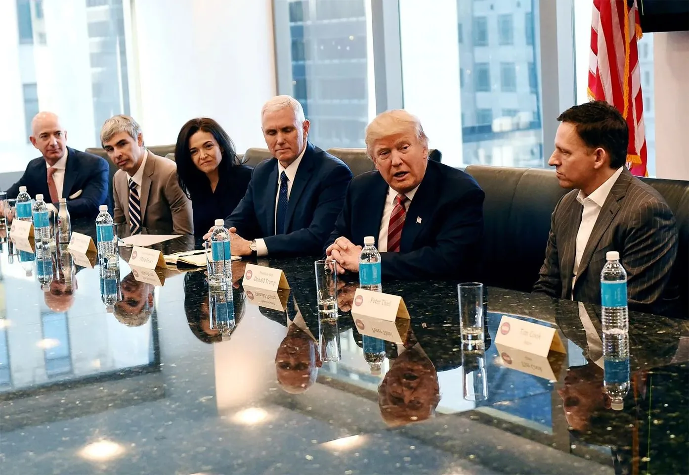 In this image, a group is gathered around a large, glossy conference table. The setting suggests a formal meeting, possibly discussing business or political matters. Jeff Bezos, identifiable by his position on the far left, is seated next to Larry Page and Sheryl Sandberg. Each person appears attentive, engaged in the conversation. Mike Pence, sitting near Donald Trump, displays a serious demeanor. Donald Trump, positioned centrally, seems to be speaking, as indicated by his open mouth and engaged posture. Peter Thiel, on the far right, listens intently. The room is well-lit, with large windows in the background allowing natural light to illuminate the scene. Each participant is dressed in formal business attire, contributing to the professional atmosphere. Name cards are placed in front of each individual, confirming their identities. Water bottles and glasses are neatly arranged on the table, adding to the organized and structured environment of the meeting.