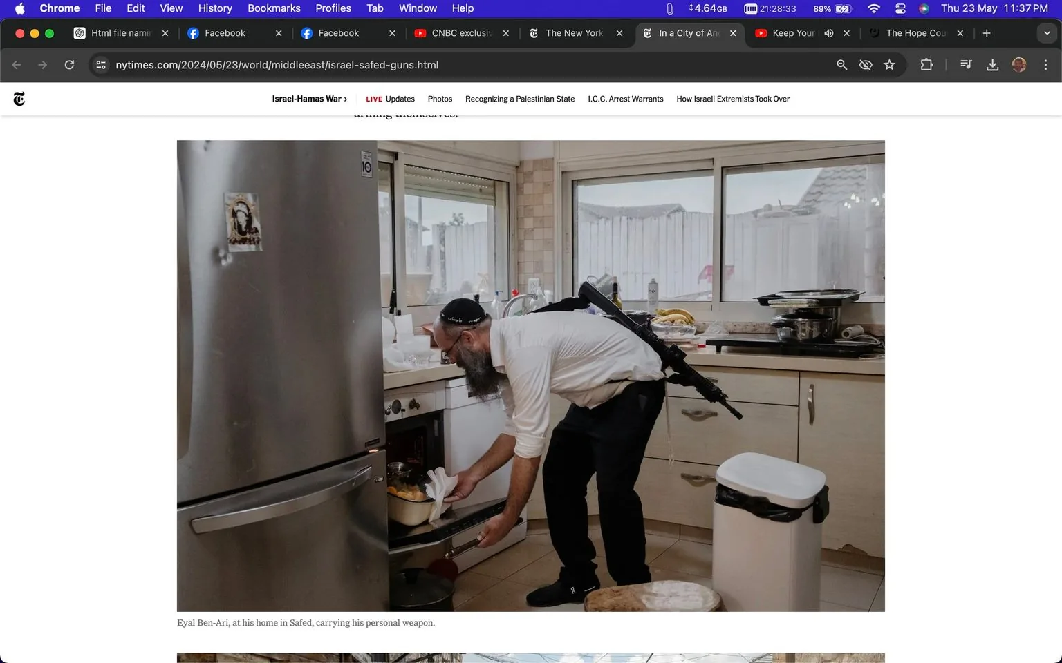 Eyal Ben-Ari is depicted in his home kitchen in Safed, engaged in a domestic activity while carrying a personal weapon slung over his back. The scene captures a moment of everyday life, showcasing a man with a long beard and wearing a white shirt, black pants, and a kippah. The kitchen is well-lit by natural light streaming through large windows in the background. The room appears tidy and functional, with a stainless steel refrigerator that has a small decoration on it, and various kitchen items like pots and dishes visible on the counters and stovetop. Eyal is bending down to place or retrieve a pot from what appears to be an oven or a cabinet, suggesting a sense of normalcy juxtaposed with the presence of the firearm. The overall composition highlights a blend of routine life and heightened personal security, reflecting the complex realities faced by residents in certain regions. The image's color palette is mostly neutral, with soft lighting contributing to the domestic atmosphere.