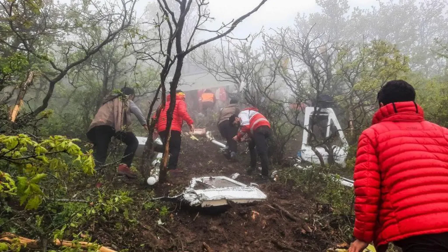 The image depicts a rescue operation on a misty, forested hillside. Several individuals in bright jackets, including red and orange, are seen moving up a muddy slope surrounded by trees. The atmosphere is foggy, adding a sense of urgency and difficulty to the scene. The terrain is rugged, with branches and debris scattered around, suggesting a recent incident. Some of the people appear to be rescuers, identifiable by their attire, while others might be assisting. Pieces of what could be wreckage are visible on the ground, hinting at a possible crash site. The overall mood is tense and focused, as the group climbs the incline, likely in an effort to reach and assist those in need. The bright colors of the jackets stand out against the muted green and brown of the forest, highlighting the rescue efforts in this challenging environment.