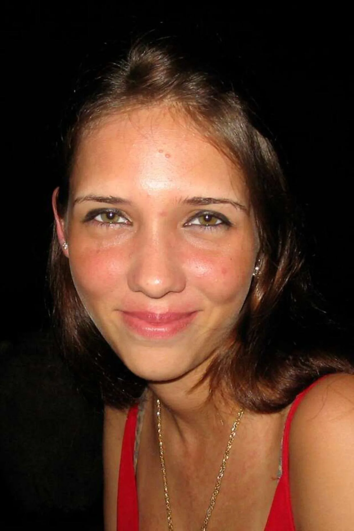 Yvonne Jonsson is featured in a close-up portrait. The image captures her in a low-light setting, suggesting it might be taken at night or in a dimly lit environment. She has a subtle smile, and her expression appears calm and content. Yvonne is wearing a red top, and a delicate gold necklace adorns her neck, adding a touch of elegance. Her hair is parted slightly off-center, and she has light makeup that highlights her natural features. The background is dark, ensuring that the focus remains on her face. The lighting creates a warm tone on her skin, emphasizing her eyes and the gentle contours of her face. The photograph has a candid feel, capturing a moment of quiet and personal reflection.