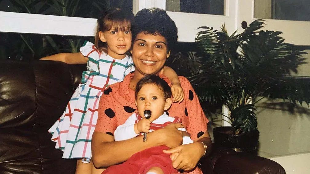 The image features a warm family moment captured indoors. A woman is seated on a brown leather couch, holding a baby in her arms. Her attire consists of a short-sleeved, patterned pink blouse, and she is smiling warmly at the camera, exuding a sense of joy and contentment. Beside her, a young girl, identified as Caroline Jonsson, is standing with her arm lovingly draped around the woman's shoulders. Caroline is dressed in a colorful, checkered dress with a white base and pink, blue, and green lines. The baby, who is sitting on the woman’s lap, is wearing a red outfit with white sleeves and is holding a spoon, looking curiously at the camera. In the background, several potted plants are visible near a large window, adding to the serene and homely atmosphere. The lighting is soft and natural, suggesting daylight filtering through the windows. The scene captures a cherished family moment, filled with affection and a sense of togetherness.