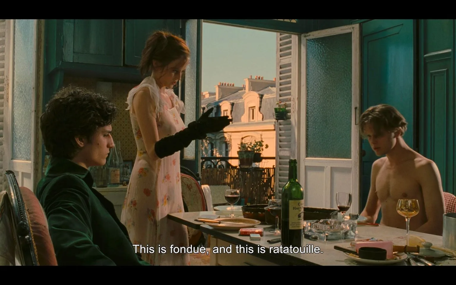The scene is from the film "The Dreamers" and features a dining setting in what appears to be a Parisian apartment. Three young adults are gathered around a table laden with wine bottles, glasses, and dishes. The atmosphere is intimate, with warm lighting suggesting evening or dusk. The woman in the center, dressed in a delicate floral dress, is holding a pair of black gloves and seems to be explaining the meal, as indicated by the subtitle, "This is fondue, and this is ratatouille." To her left, a man with curly hair sits back in his chair, wearing a dark jacket, while another man, shirtless, is seated to her right, appearing contemplative or engaged in thought. The background showcases an open window with a view of classic Parisian architecture, adding to the film's European ambiance. The scene captures a moment of casual conversation and camaraderie, with an artistic composition that emphasizes the characters' interactions and the setting's elegance.