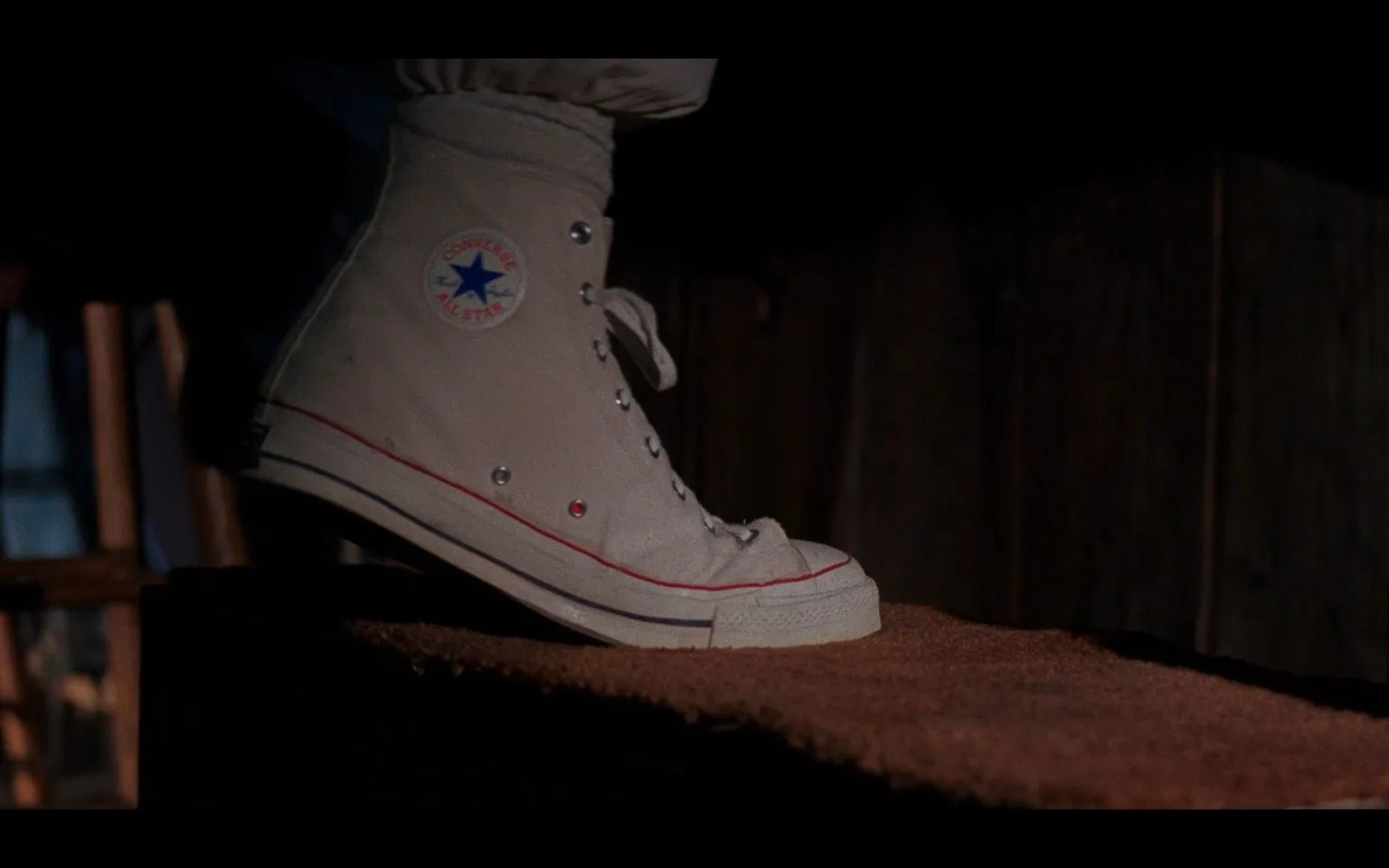 The image is a close-up shot of a single foot wearing a classic Converse shoe, specifically a white high-top Chuck Taylor All Star. The shoe is prominently displayed with the iconic circular logo visible, featuring a blue star and the text "Converse All Star" in red. The shoe's design includes metal eyelets for the laces, a red line along the sole, and a blue stripe beneath it, typical of this classic footwear style. The fabric appears slightly worn, suggesting usage. The lighting is low, indicating a nighttime setting or a dimly lit interior. The background is blurred but seems to include wooden elements, hinting at a rustic or unfinished space. The mood is subtle and focuses on the nostalgic and timeless appeal of the Converse brand. There is no visible text or title information in the image.