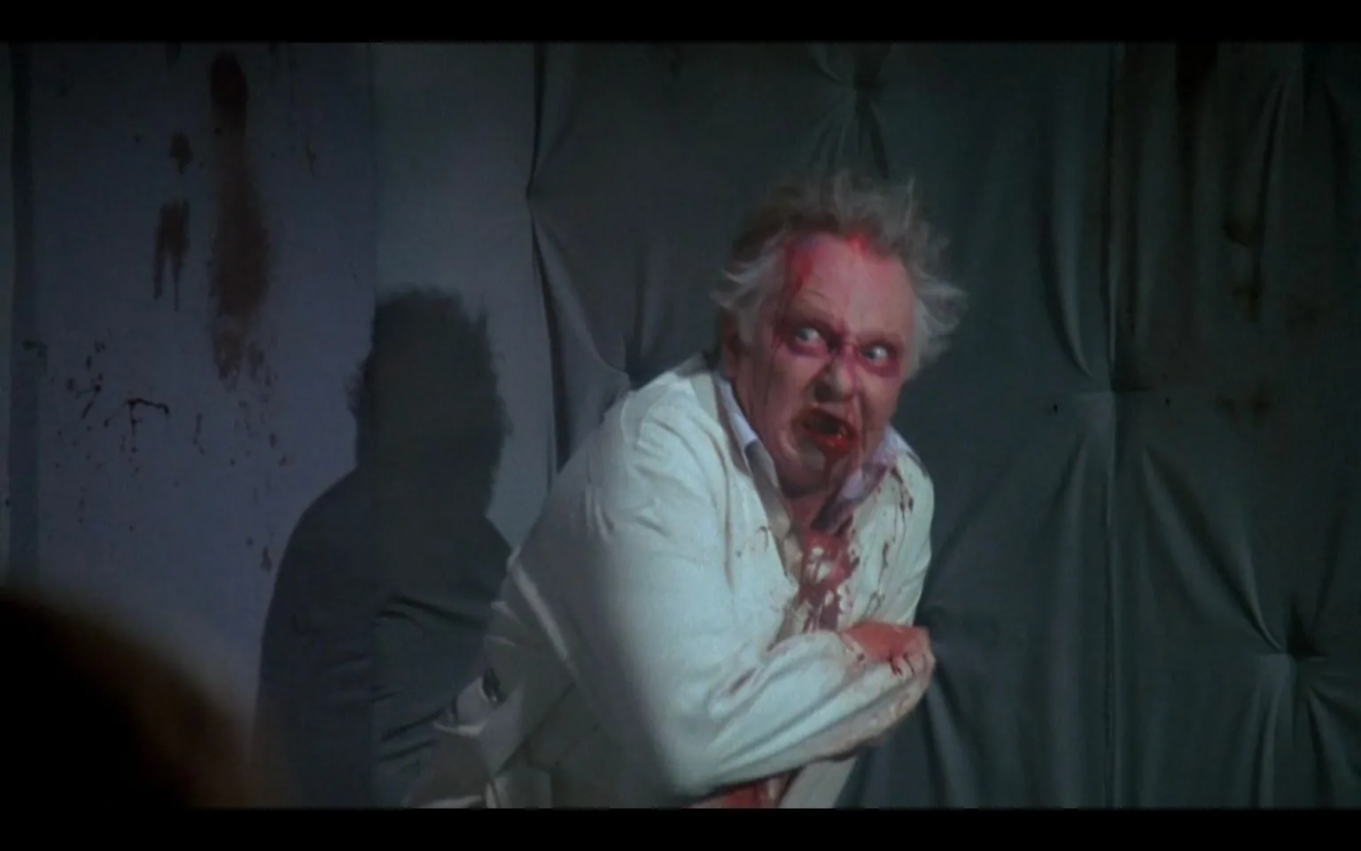 The image depicts a dramatic scene set in what appears to be a psychiatric ward or padded cell. A man with disheveled hair and a bloodied face is wearing a white, possibly hospital-issue, outfit. The expression on his face is intense and menacing, with wide eyes and a grimace that suggests anger or madness. Blood stains are visible on his clothing and smeared across his face, adding to the horror-like atmosphere.
The background features padded walls, which are typically associated with psychiatric facilities, enhancing the sense of confinement and tension. Lighting in the scene casts a shadow of the man on the wall, adding an ominous element to the composition. There are splatters of blood on the wall, contributing to the unsettling and chaotic mood of the image. Overall, the scene is vividly intense, capturing a moment of violent emotion and psychological disturbance.