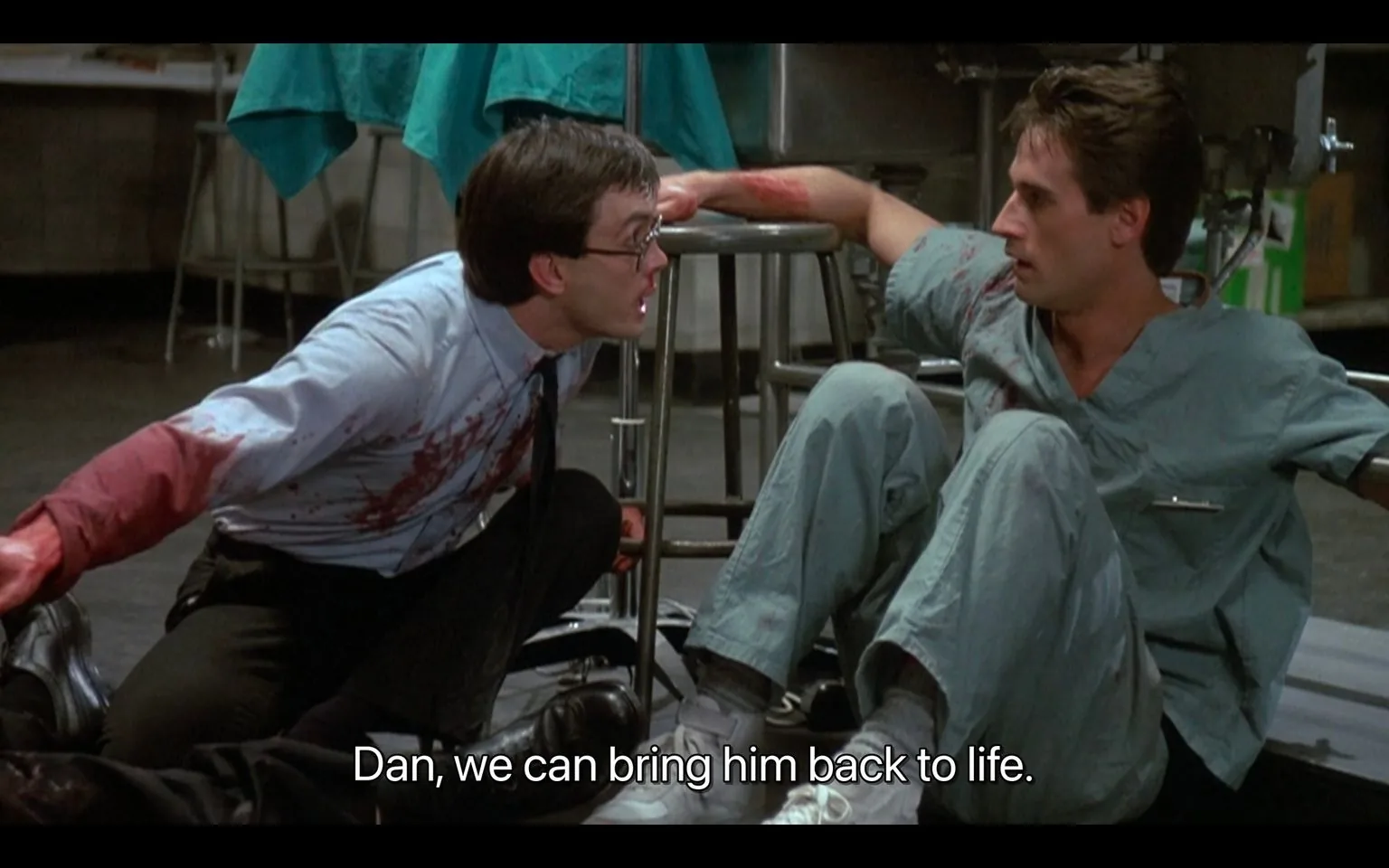 The scene is from the film "Re-Animator," capturing a tense moment in what appears to be a laboratory setting. Two men are central to the frame, with one wearing a blood-stained white shirt and glasses, leaning towards the other, who is in scrubs. The man in the shirt, with a determined and urgent expression, speaks passionately to the other, emphasizing the possibility of reviving someone. The man in scrubs is seated on the floor, leaning against a stool, appearing shocked or contemplative as he listens.
The background consists of medical equipment and a teal-colored sheet draped over a surgical table, reinforcing the setting's clinical atmosphere. The lighting is stark and bright, highlighting the blood on the first man's shirt and the intensity of the situation. The caption, "Dan, we can bring him back to life," suggests a pivotal moment in the narrative, hinting at an experimental or supernatural attempt at resurrection. The frame captures the suspense and horror elements characteristic of the film.