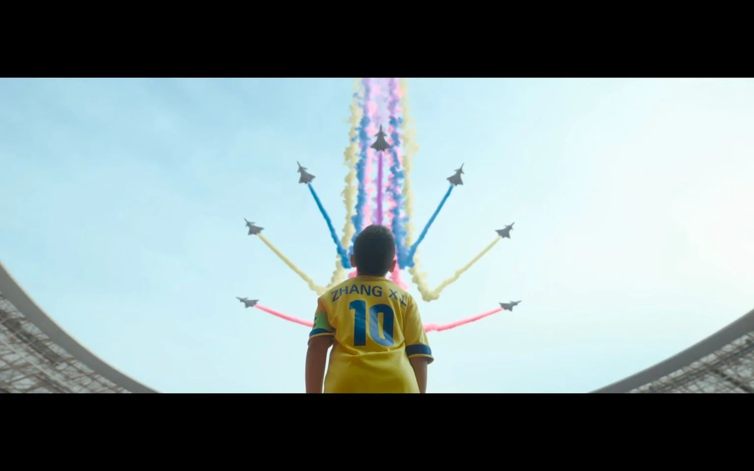 In this striking image, a young boy stands in a stadium, looking up at a captivating airshow above. The boy is wearing a yellow sports jersey with the name "ZHANG XI" and the number "10" on the back, suggesting he might be a soccer enthusiast or player. The stadium is large, with the roof partially visible around the edges of the image, framing the scene above.
The sky is filled with jets streaming colored smoke trails in vibrant hues of blue, yellow, and red. The jets are performing an aerial display, creating a dramatic spectacle against the clear, bright sky. The lighting is natural and emphasizes the vivid colors of the smoke trails, adding to the scene's energy and excitement. The boy's posture is one of awe and wonder, capturing the spirit of the moment.
The overall composition centers on the boy, with the jets forming a dynamic pattern that draws the viewer's eye upward, symbolizing ambition and dreams. The image conveys a sense of inspiration and the thrill of witnessing a grand event.
- Key OCR Lines: "ZHANG XI 10"