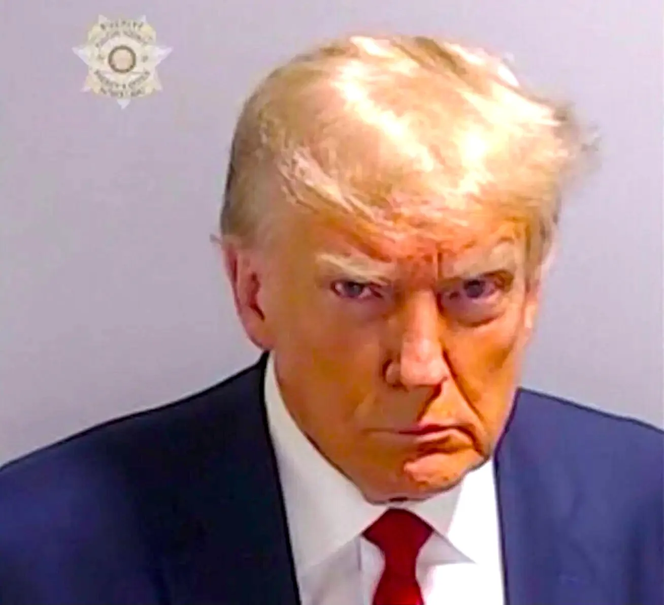 Donald Trump is depicted in this image, which appears to be a mugshot. The setting is indoors with a plain, neutral background. Donald Trump is wearing a dark suit jacket, a white shirt, and a bright red tie. His expression is serious and focused, with a direct gaze at the camera. The lighting is even and illuminates his face and upper body. In the top left corner, there is a sheriff's badge, suggesting the context of a law enforcement setting. The image has a formal and official tone, characteristic of legal documentation. The colors are vivid, especially noticeable in the contrast between his attire and the background. Overall, the image conveys a sense of formality and gravity.