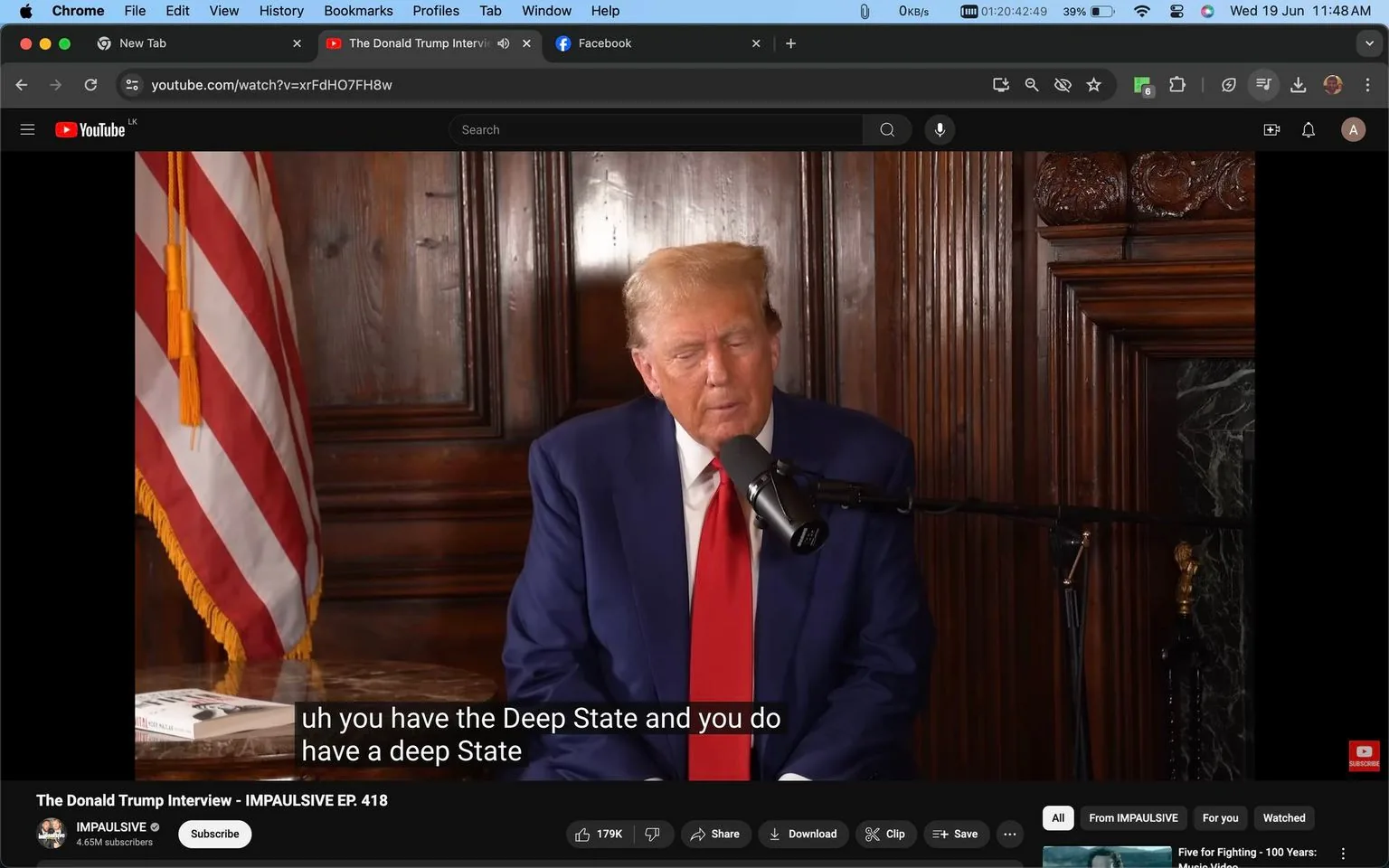 The screenshot captures a scene from a YouTube video titled "The Donald Trump Interview - IMPAULSIVE EP. 418" on the "IMPAULSIVE" channel. The setting is indoors, featuring a wooden panel background and an American flag to the left side of the frame, indicating a formal or official setting. In the center, a man, identified by the hint as Trump, is seated and speaking into a microphone. He is dressed in a navy blue suit with a red tie, which are typical colors associated with patriotism in the United States. His expression is focused and serious, suggesting a discussion of significant political topics. The lighting is warm and even, highlighting the wood textures behind him. A small table with a book is visible in the left foreground. The subtitles at the bottom of the screen feature the text, "uh you have the Deep State and you do have a deep State," indicating the subject matter being discussed in the interview. The layout suggests a professional and polished production intended for a broad audience.
