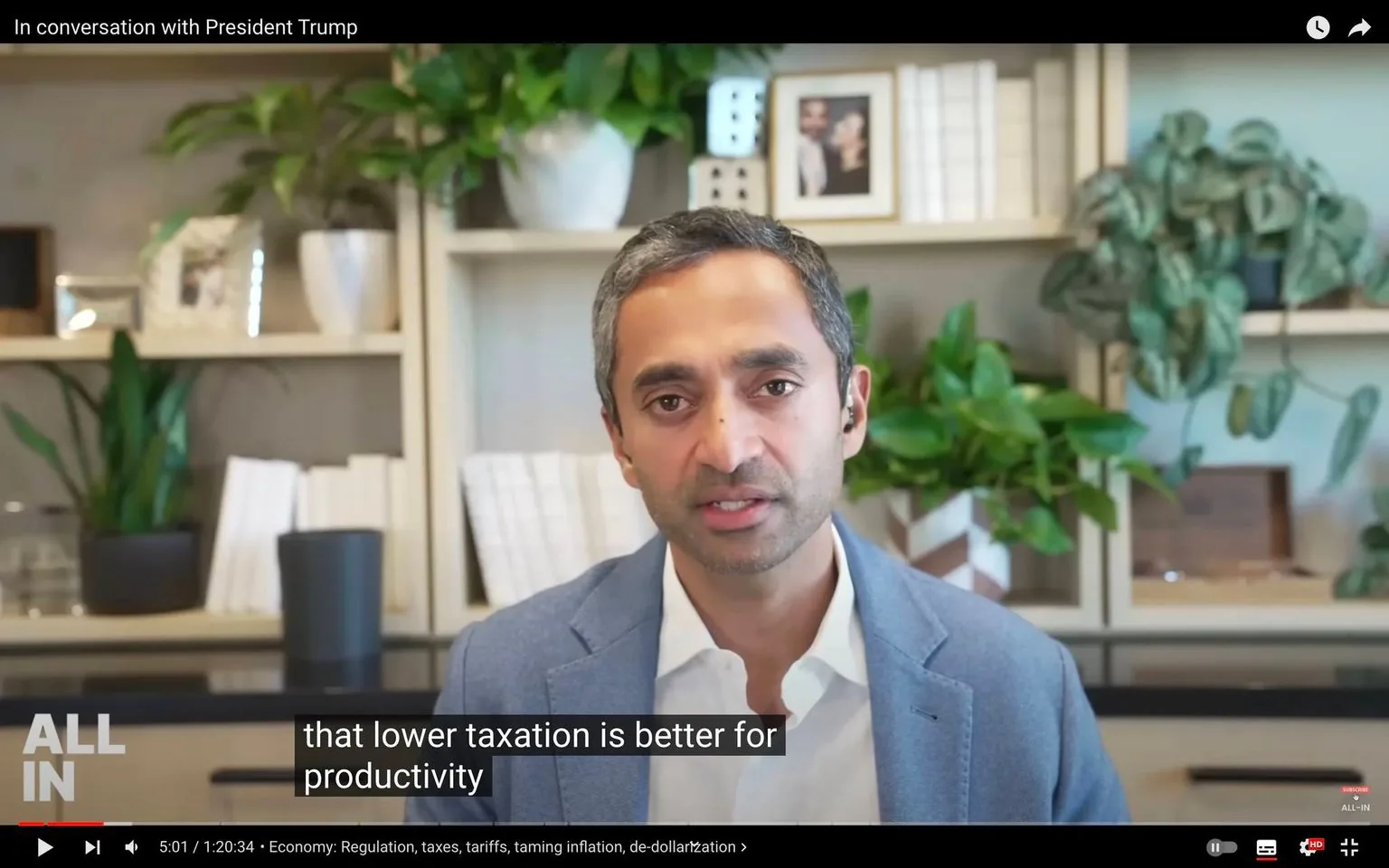 The image is a video frame from a YouTube channel called "ALL IN," featuring a person in an interview setting. The video title is "In conversation with President Trump," suggesting a discussion related to economic topics. The individual in the frame is speaking directly to the camera, possibly via video call, and is wearing a blue blazer over a white shirt. The background reveals a well-organized office or study, adorned with shelves containing books, potted plants, and framed photographs, adding a personal touch to the setting.
The on-screen captions indicate the discussion is about the impact of lower taxation on productivity, hinting at themes of economic policy and regulation. The video frame is well-lit, conveying a professional and formal atmosphere. The person appears focused and engaged, contributing to a serious yet conversational tone. The surrounding plants and books soften the office environment, balancing professionalism with a touch of warmth and familiarity.