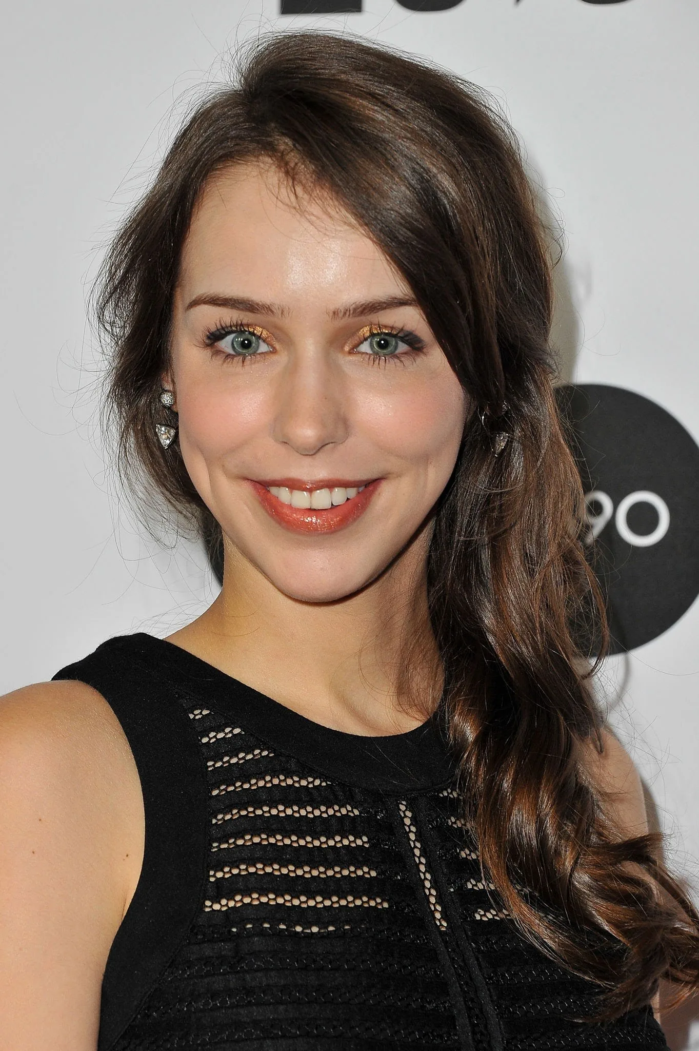 Stefanie Joosten is featured in this portrait-style image. She is indoors, likely at an event, given the professional backdrop. She has long brown hair styled in soft waves cascading over one shoulder. Her makeup is understated, highlighting her eyes with subtle gold eyeshadow and a natural lip color. Stefanie is wearing a sleeveless black top with intricate mesh detailing, giving a chic and stylish appearance. Her expression is warm and welcoming, with a bright smile that adds to the positive mood of the image. The lighting is soft, enhancing the natural tones of her skin and hair. The background includes a partially visible logo, suggesting a formal or media event setting.