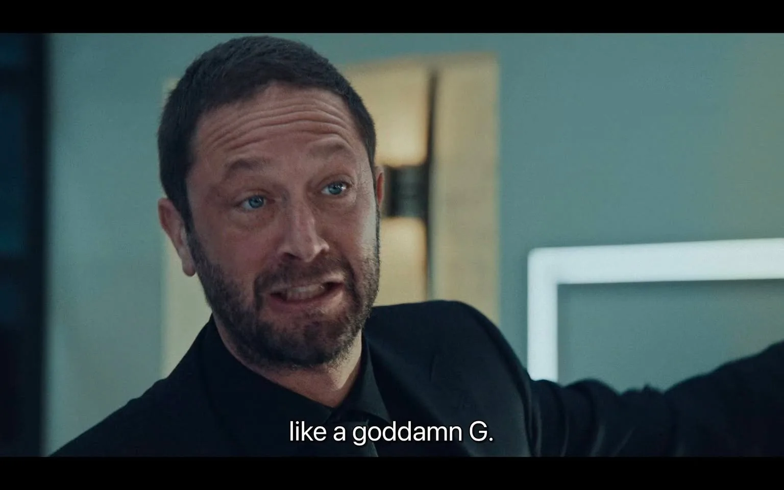 The image is a screenshot from a scene featuring a man with a beard, wearing a black suit. The frame is captured indoors, likely during a conversation. The man's expression is intense, and he appears to be in the middle of speaking emphatically. The lighting is bright and focused, creating a professional or serious atmosphere. In the background, there is a blurred section of the room with a modern, minimalist design. The subtitle at the bottom reads, "like a goddamn G." This suggests that the dialogue might involve themes of confidence or assertiveness. The composition centers on the man's face and upper body, emphasizing his expression and hand gesture as part of the conversation.