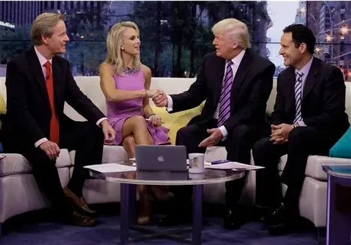 In the image, four individuals are seated on a light-colored couch, engaging in a conversation typical of a talk show setting. Elisabeth Hasselbeck is among them, wearing a sleeveless, purple dress. She is positioned second from the left and is shaking hands with a man in a dark suit and striped tie, who is likely Donald Trump, as indicated by the hint. The two other men are also dressed in dark suits and ties, contributing to the formal atmosphere of the scene.
The setting appears professional and polished, with a cityscape visible through large windows in the background, suggesting a high-rise location. A small table in front of them holds a laptop and some papers, reinforcing the talk show or interview scenario. The lighting is bright and even, highlighting the participants and creating a lively, engaging environment. The expressions and body language suggest a friendly and conversational interaction among the group.