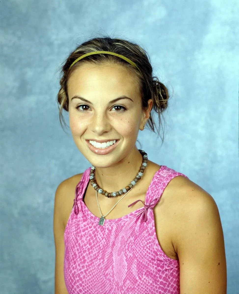 Elisabeth Hasselbeck is depicted in this studio portrait. She is wearing a pink sleeveless top with a textured pattern resembling snakeskin. Her hair is styled with a headband, and it is pulled back in a casual updo. Elisabeth is accessorized with a beaded necklace and a small pendant necklace. The background is a light blue, textured surface, giving the image a calm and neutral setting. Her expression is cheerful, with a broad smile that conveys a friendly and approachable demeanor. The lighting is soft and even, highlighting her features without any harsh shadows. This image likely captures her in a professional or promotional context, aiming to present her in a positive and vibrant light.